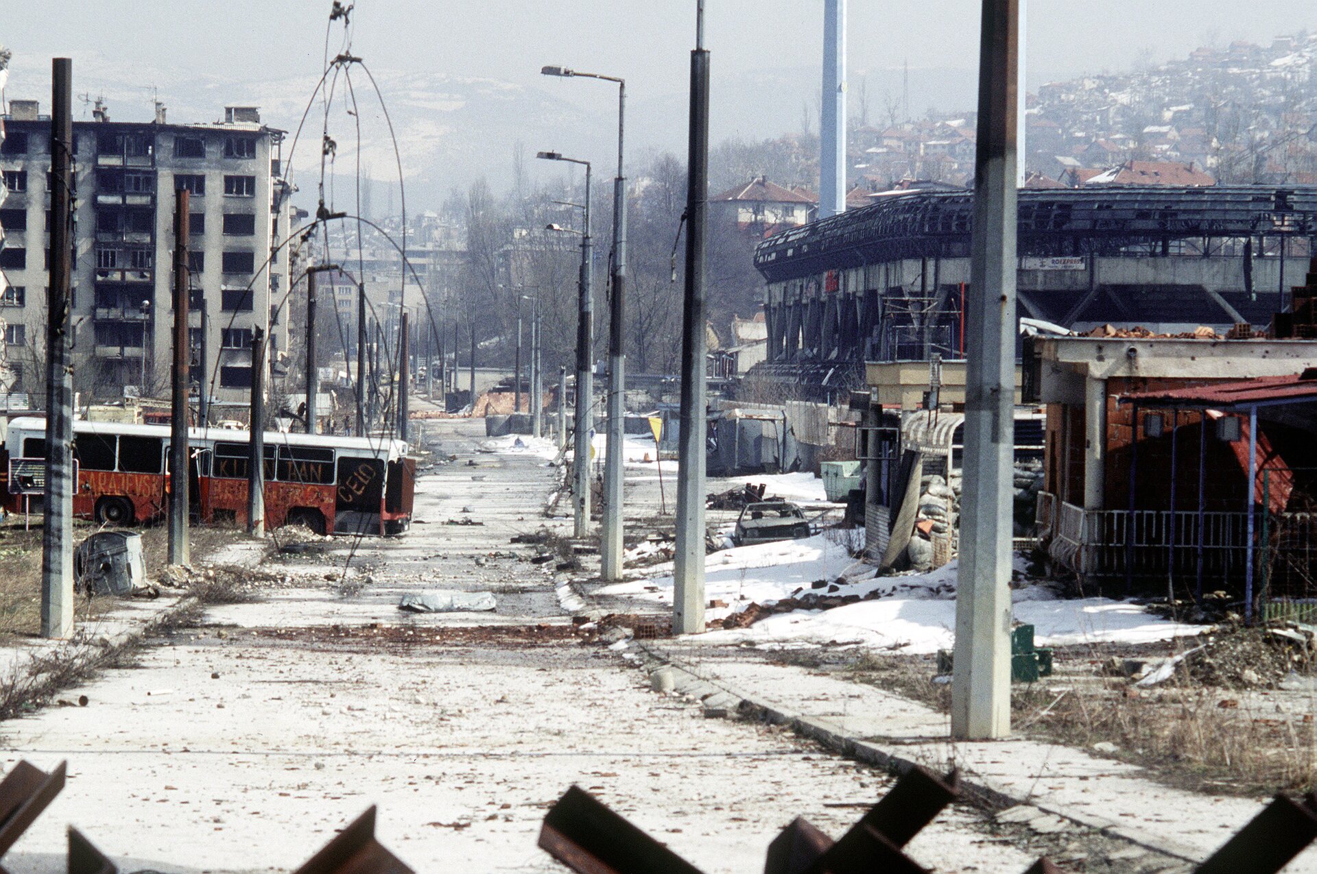 Zdjęcie przedstawia Sarajewo po wojnie w latach 1992 – 1996. Środek zdjęcie zajmuje asfaltowa ulica biegnąca wzdłuż centrum miasta. Po obu stronach wysokie betonowe latarnie. W głębi zdjęcia zniszczony spalony autobus. Autobus stoi w poprzek . Po obu stronach ulicy zniszczone zabudowania. Ulica, chodniki, pobocza i budynki są pokryte cienką warstwą śniegu. W głębi zdjęcia, za miastem, budynki na zboczu wzgórza. Wzgórze znajduje się po prawej stronie miasta. Dachy budynków pokryte białą warstwą śniegu.