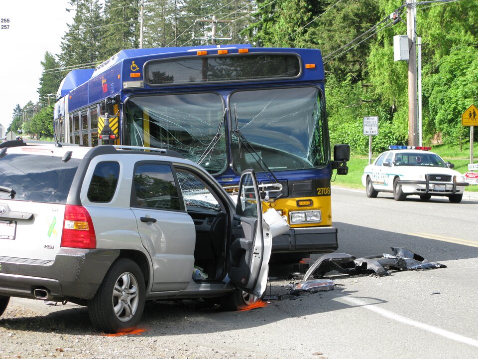 Zdjęcie przedstawia wypadek drogowy z udziałem autobusu. Słoneczny dzień. Autobus i samochód osobowy znajdują się na asfaltowej dwupasmowej jezdni. Niebiesko żółty autobus ustawiony przodem do obserwatora zdjęcia. Biały samochód osobowy bokiem do obserwatora zdjęcia. Przednie szyby autobusu są pęknięte poprzecznie, ale nadal na miejscu. Samochód osobowy, który zderzył się z autobusem czołowo ma całkowicie zniszczony przód. Na asfalcie kawałki czarnego zderzaka samochodu. Drzwi pasażera otwarte. Widoczne w kadrze koła samochodów mają oznaczone pomarańczową farbą w spreju miejsca, w których stykają się z asfaltem. W tle, na prawo, samochód policyjny. Na dachu samochodu policyjnego niebieskie i czerwone prostokątne światła.