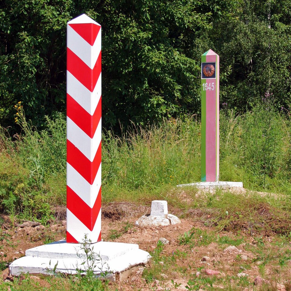 Fotografia prezentuje dwa znaki graniczne. Na pierwszym planie słupek pomalowany w poziome biało-czerwone pasy, na drugim planie słupek pomalowany w pionowe fioletowo-zielone pasy z numerem i emblematem na szczycie.