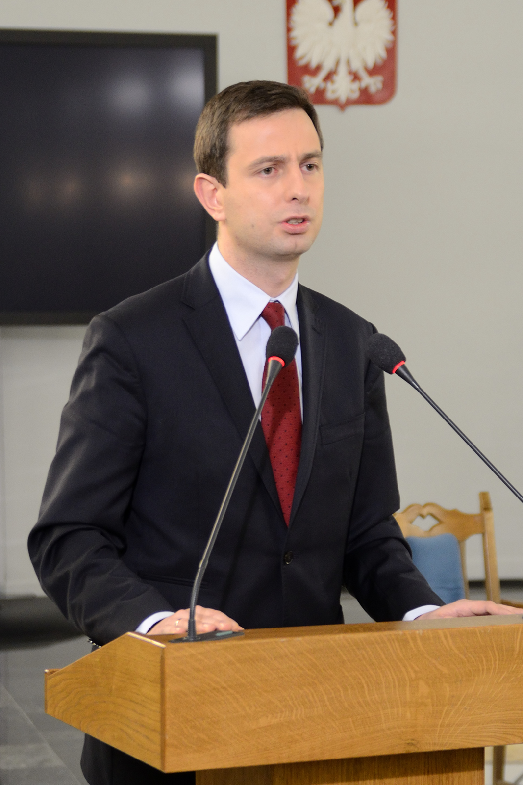 Kolorowe zdjęcie przedstawia Władysława Kosiniak-Kamysza. Mężczyzna stoi przodem do obserwatora zdjęcia. Mężczyzna w wieku około 40 lat. Włosy ciemne gładko zaczesane na lewą stronę. Czoło wysokie, gładkie. Brwi jasne, gęste. Oczy ciemne, małe. Nos wąski, długi. Usta wąskie. Twarz owalna, gładko ogolona. Uszy duże. Ubrany w czarną marynarkę, białą koszulę i ciemno czerwony krawat.