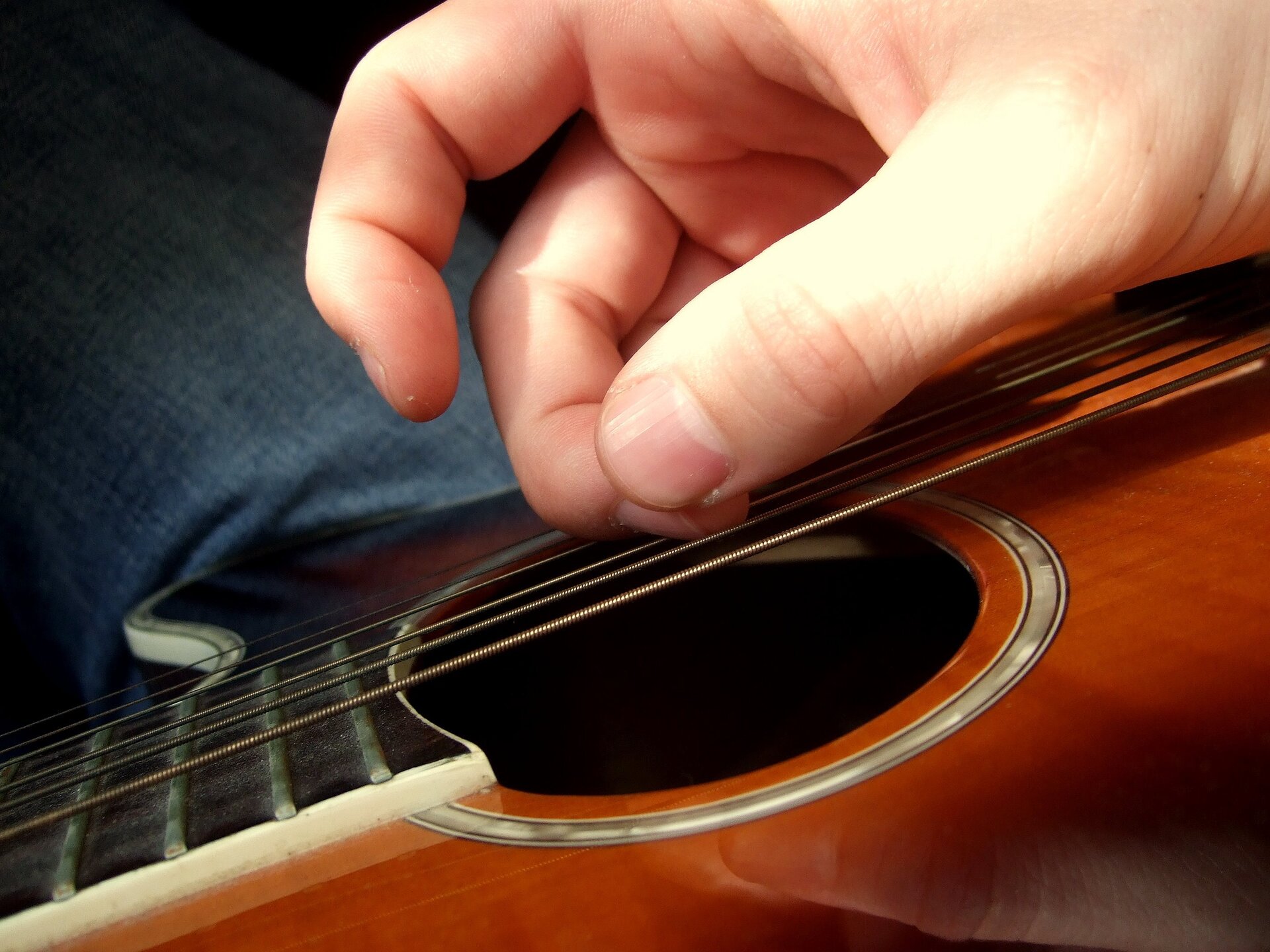 Zdjęcie przedstawia zbliżenie palców prawej ręki szarpiących struny gitary akustycznej z perspektywy osoby grającej na tę gitarze. W tle materiał spodni, gitara oparta na nodze grającego. W kadrze mieści się centralna część pudła oraz niewielki fragment gryfu.