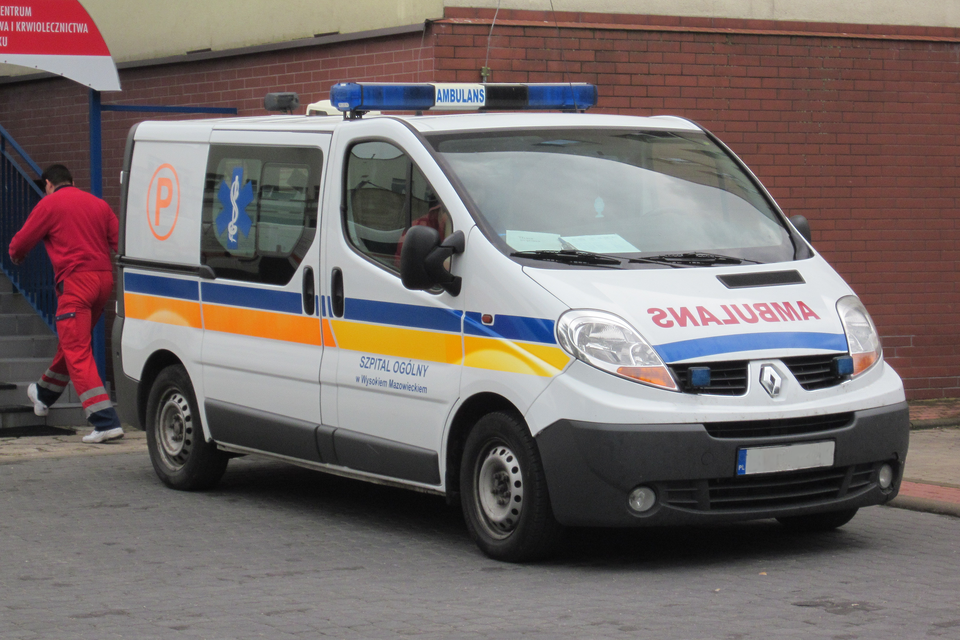 Fotografia prezentuje białą karetkę pogotowia ratunkowego stojącą przed budynkiem. Z przodu karetki, pod przednia szybą i między światłami, znajduje się czerwony napis SNALUBMA, który jest lustrzanym odbiciem napisu AMBULANS.