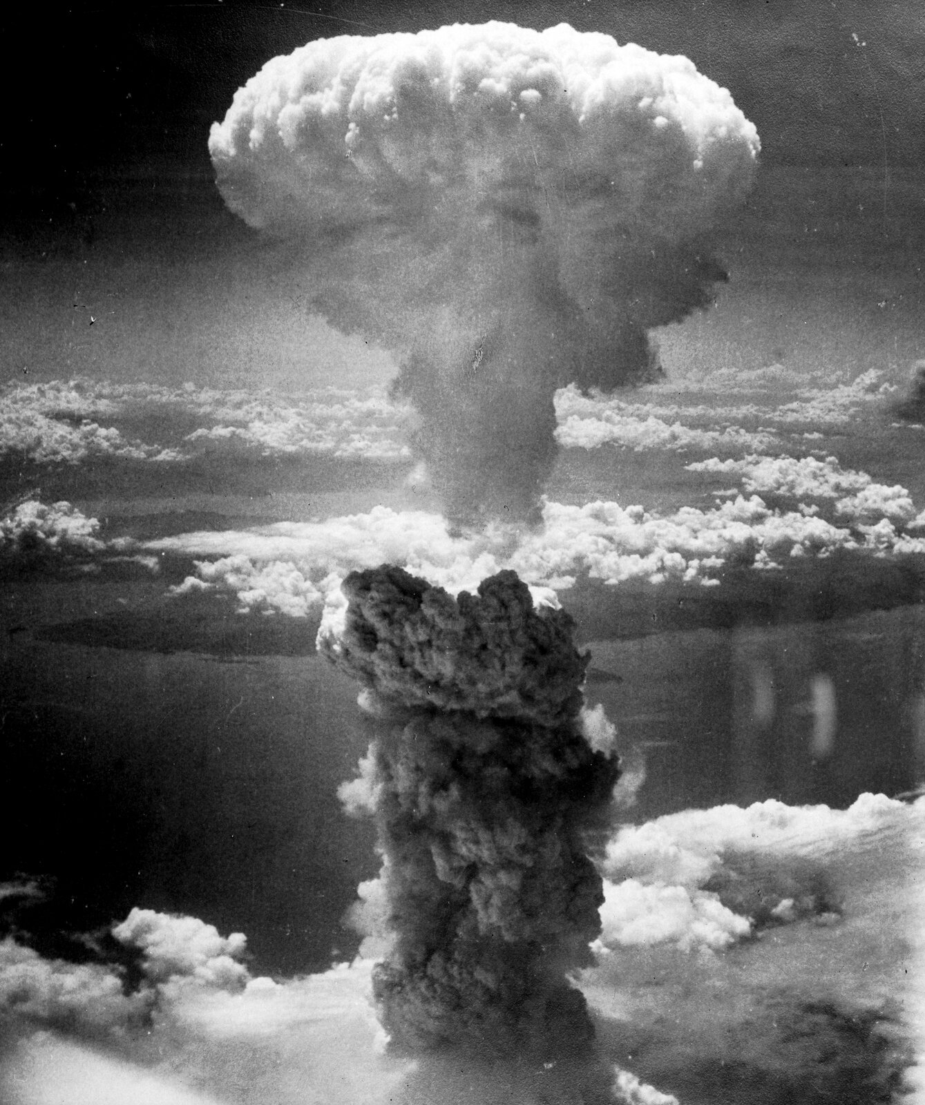 Historyczne zdjęcie czarnobiałe wykonane z pokładu bombowca B-29 przedstawia efekt wybuchu bomby atomowej Fat Man nad Nagasaki. Na pierwszym planie widoczny jest obłok w kształcie grzyba u dołu czarny, a u góry biały. Daleko poniżej chmury, morze i wyspy. Obszar z którego wyrasta czarny słup to ląd częściowo przesłonięty chmurami. Brak widocznych szczegółów ukształtowania terenu.