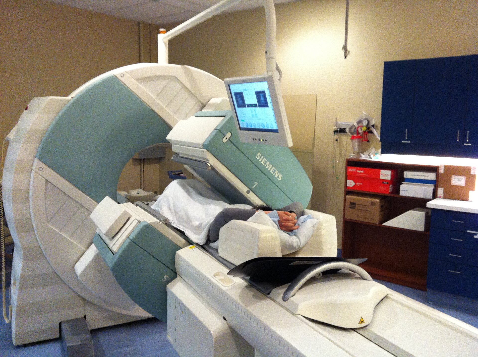 Na zdjęciu wnętrze gabinetu diagnostycznego. Na środku tomograf. Duże koło z otworem pośrodku. Na ruchomej części tomografu leży pacjent. Na monitorze obraz diagnozowanego narządu pacjenta. Z prawej strony inne wyposażenie gabinetu, regały z segregatorami.