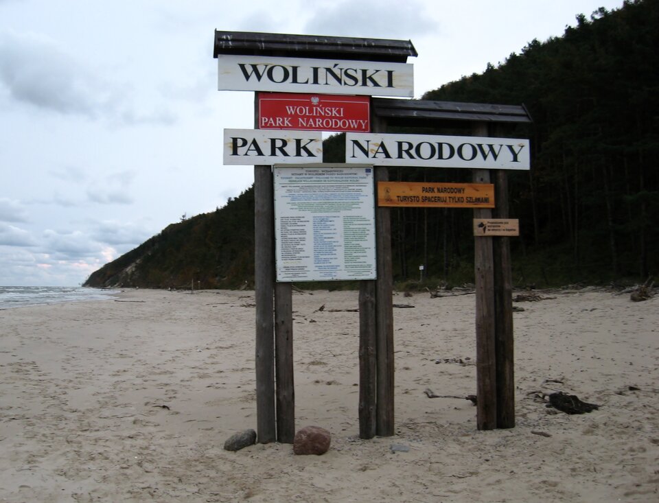 Na zdjęciu tablica informacyjna na plaży. Drewniane słupki, daszek. Napisy i informacje.