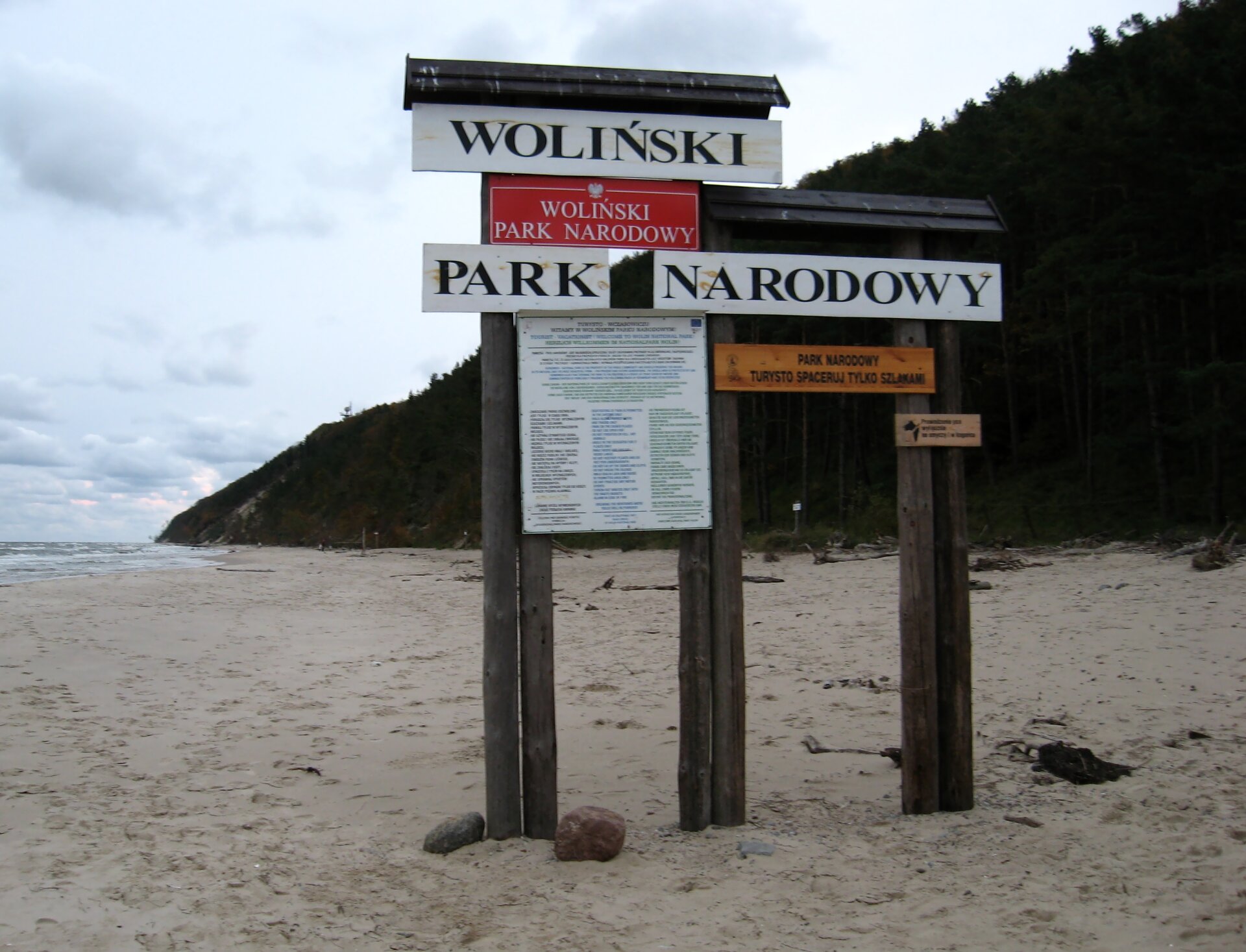 Na zdjęciu tablica informacyjna na plaży. Drewniane słupki, daszek. Napisy i informacje.
