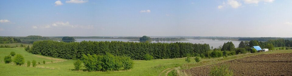 Fotografia przedstawia krajobraz rolniczy. Na pierwszym planie z lewej łąka z zaroślami, z prawej pole uprawne, droga i zabudowania. W centrum za ciemno zielonym lasem rozciąga się szarobłękitny pas jeziora. Na nim kępy drzew na wyspach. To Jezioro Siedmiu Wysp albo Oświn, jeden z obszarów Konwencji Ramsar w Polsce.