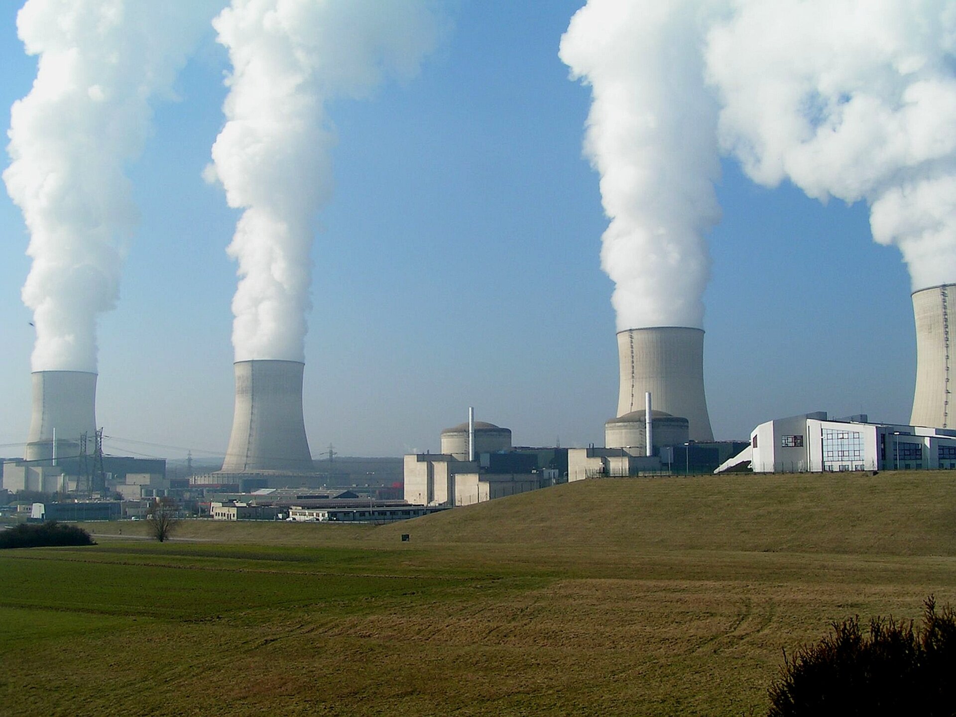 Kolorowe zdjęcie przedstawia budynki i chłodnie elektrowni atomowej w Cattenom. Słoneczny dzień. Na pierwszym planie szeroki płaski teren pokryty zielono-żółtą krótką trawą. Chłodnie elektorowi mieszczą się w głębie. Ich kształt można określić jako cylindryczny. Biała gęsta para wydobywa się z chłodni i pionowo wzbija się w górę. Pomiędzy chłodniami niskie zabudowania elektrowni.