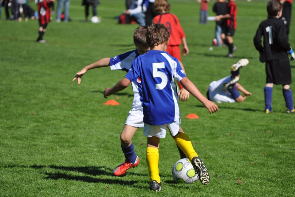 Fotografia przedstawia chłopców grających na boisku w piłkę nożną. Na pierwszym planie dwaj chłopcy w strojach sportowych jednej drużyny, w profesjonalnym obuwiu do gry w piłkę nożną, podają sobie piłkę, kopiąc ją po murawie. Na dalszym planie kilkunastu chłopców w strojach sportowych różnych drużyn ćwiczy.