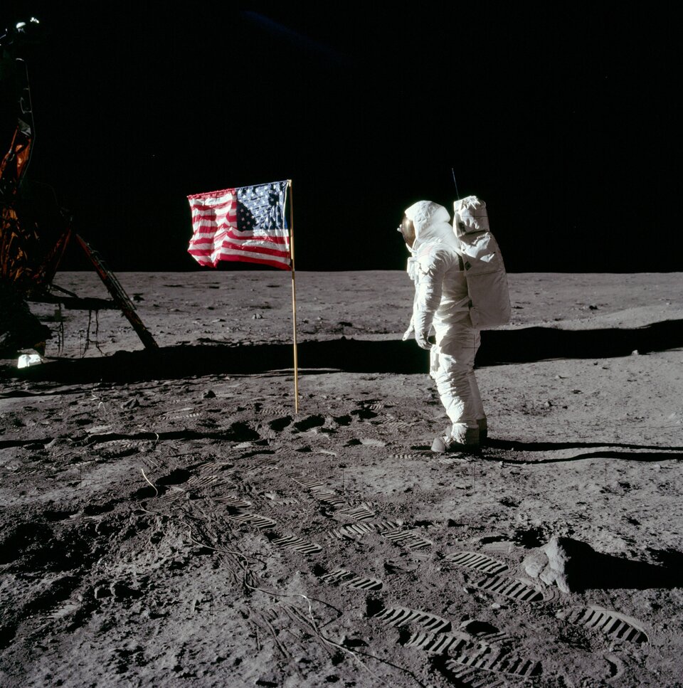 Zdjęcie ze zbiorów Amerykańskiej Agencji Aeronautyki, przedstawiające misję Apollo 11 i pierwsze lądowanie człowieka na Księżycu. Kadr kwadratowy, po lewej stronie widoczny fragment lądownika, centrum kadru zajmują flaga USA oraz salutujący jej astronauta Buzz Aldrin. W dolnej części kadru wyraźne ślady astronautów na powierzchni Księżyca.