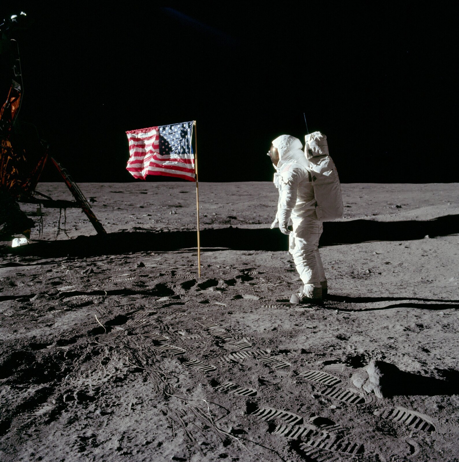 Zdjęcie ze zbiorów Amerykańskiej Agencji Aeronautyki, przedstawiające misję Apollo 11 i pierwsze lądowanie człowieka na Księżycu. Kadr kwadratowy, po lewej stronie widoczny fragment lądownika, centrum kadru zajmują flaga USA oraz salutujący jej astronauta Buzz Aldrin. W dolnej części kadru wyraźne ślady astronautów na powierzchni Księżyca.