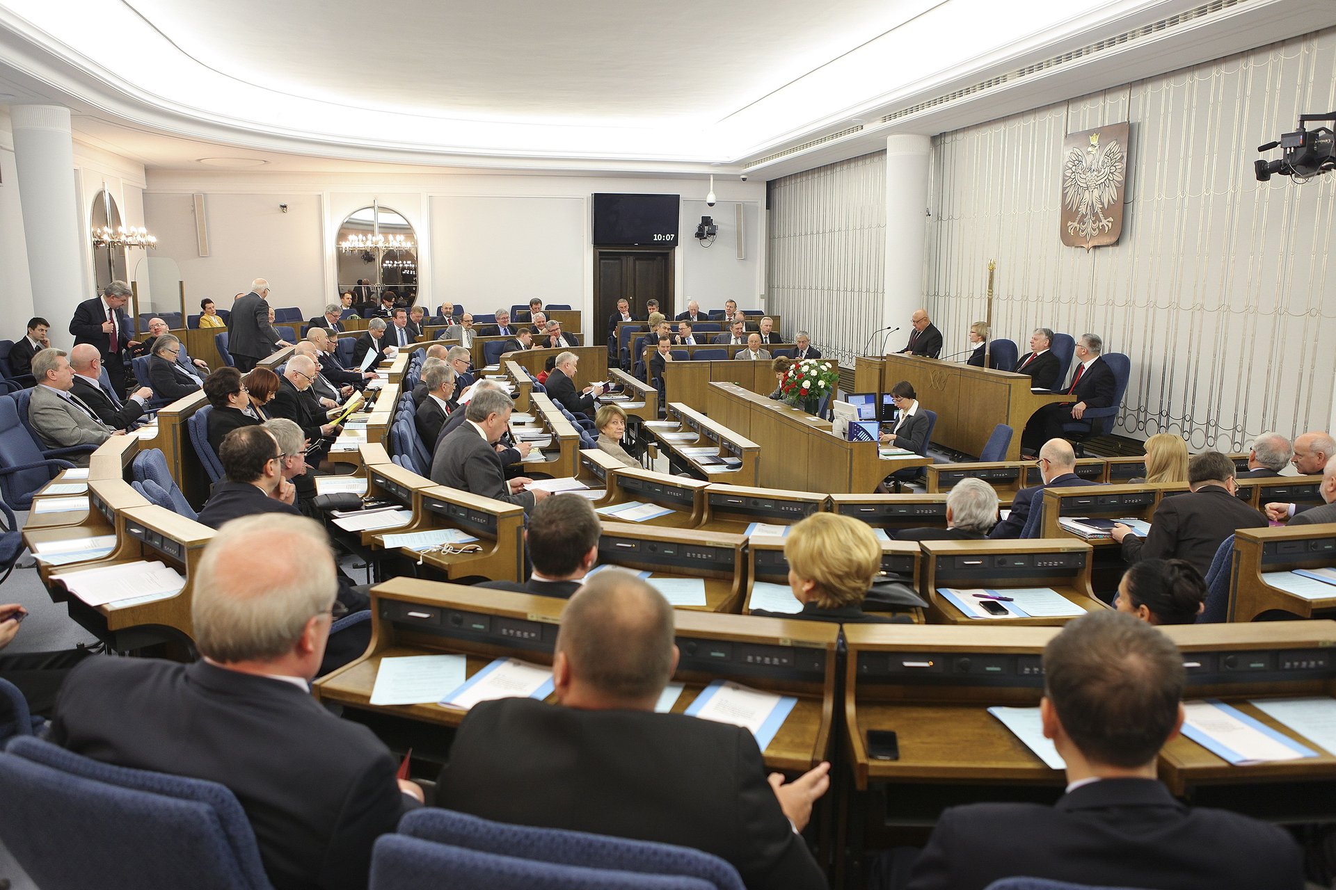 Kolorowe zdjęcie sali Senatu RP. Po prawej stronie biała ściana z zawieszonym godłem Polski. Biały orzeł w koronie na czerwonym tle. Naprzeciw godła 4 rzędy ławek z miejscami dla członków senatu. Ławy w kształcie elipsy. W głębi duże okrągłe lustro. Członkowie senatu zasiadają w swoich miejscach. Na pulpitach białe kartki.