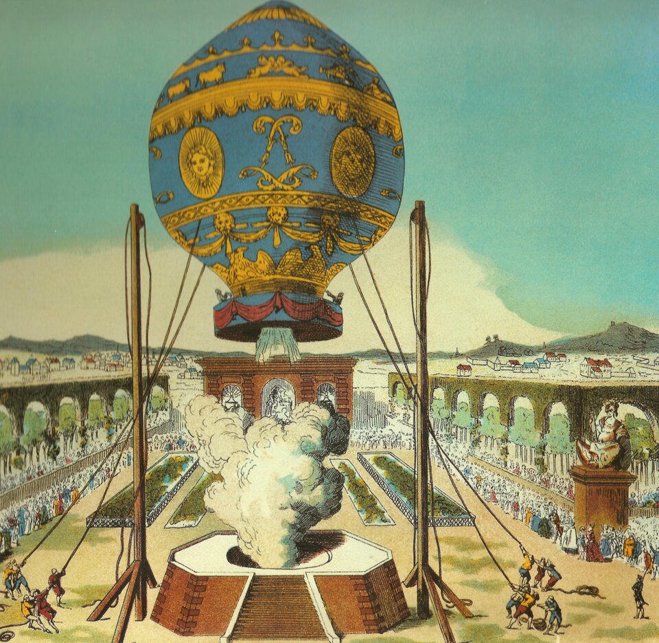 Ilustracja przedstawia wielki balon powietrzny. Niebieski z żółtymi malowidłami. Balon unosi się. Przy pomocy lin przytrzymywany jest przez kilkoro ludzi znajdujących się na dole. Pod balonem widoczne biało-szare kłęby mgły lub dymu. W tle tłumy ludzi.