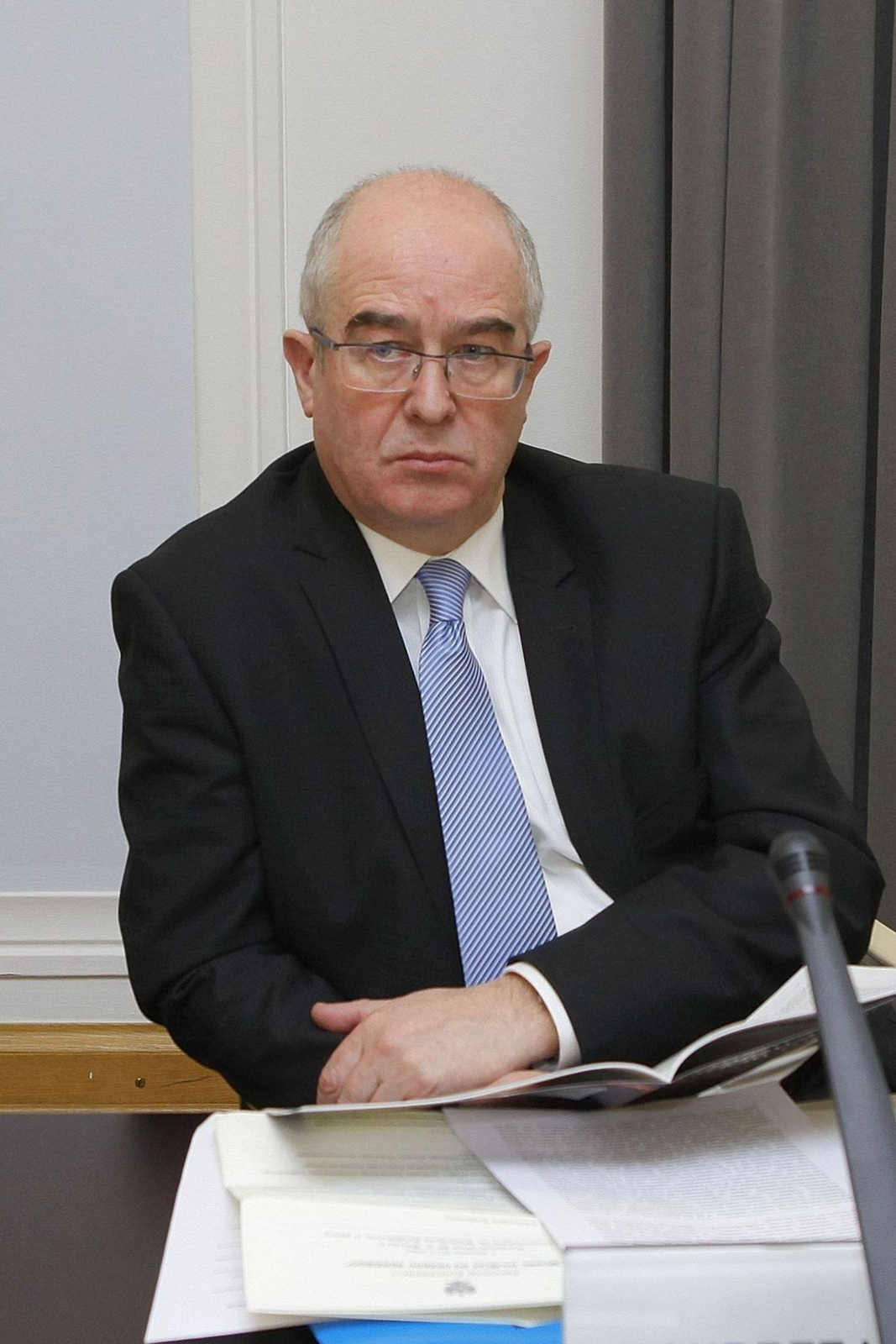 Kolorowe zdjęcie Andrzeja Seremeta. Mężczyzna, w wieku około 55 lat, siedzi przy stole. Twarz okrągła, policzki pulchne. Siwe włosy gładko zaczesane nad uszami. Na górze głowy łysina. Czoło wysokie z poziomymi zmarszczkami. Brwi ciemne, gęste. Oczy małe. Nos duży. Usta wąskie. Okulary w metalowych oprawkach. Szyja szeroka. Ubrany w czarną marynarkę, białą koszulę i błękitny krawat.