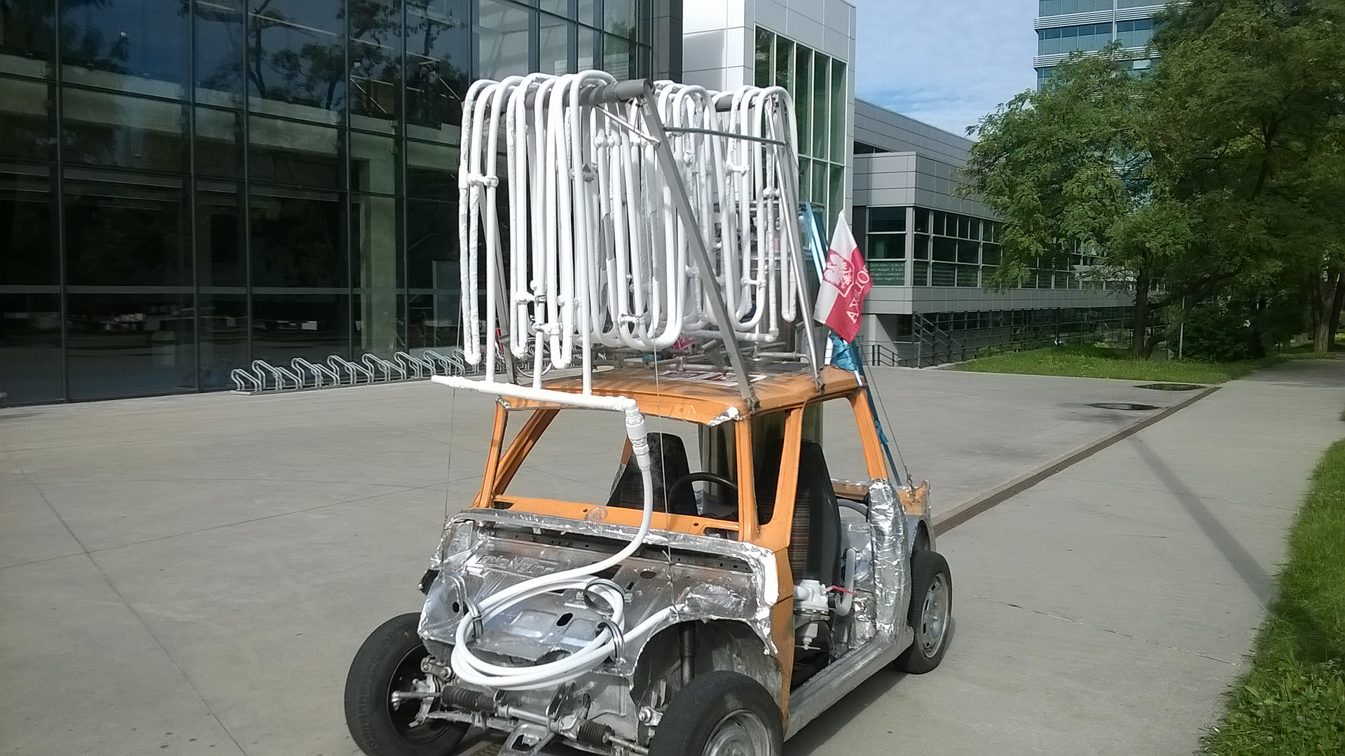 Zdjęcie przedstawia pojazd zbudowany na bazie szkieletu Fiata 126 p ze skomplikowanym układem białych rurek na dachu. Gruby przewód od tych rurek biegnie do układu znajdującego się gdzieś pod srebrną częścią obudowy z ocynkowanej stali lub aluminum. Pojazd ma dwa miejsca siedzące, jest pozbawiony szyb i drzwi. Stoi na betonowej płycie przed oszklonym budynkiem uczelni.