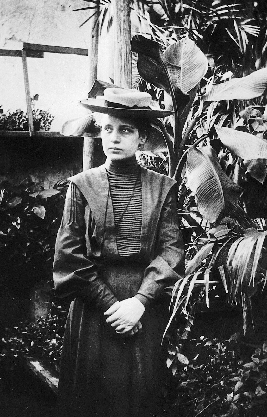 Zdjęcie przedstawia Lisę Meitner. Fotografia czarno-biała. Na zdjęciu młoda, szczupła kobieta, w wieku ok. 30 lat, ubrana w długą sukienkę z długim rękawem. Na głowie kapelusz. Dłonie złożone przed sobą, jedna na drugiej. Oczy duże, nos prosty, usta średnie, policzki pełne.