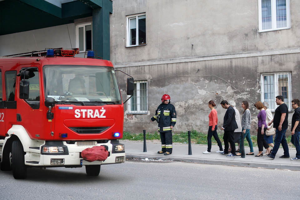 Zdjęcie przedstawia ewakuację grupy ludzi z budynku. Ulica. Słoneczny dzień. Na pierwszym planie, po lewej stronie, czerwony wóz strażacki. Wóz strażacki skierowany w prawo. Na prawo od wozu, chodnik. Wzdłuż chodnika prowadzeni są mieszkańcy. Z przodu grupy strażnik. Strażnik ubrany w kombinezon ochronny oraz czerwony kask. Za ludźmi, w tle zdjęcia, wysoki szary budynek.
