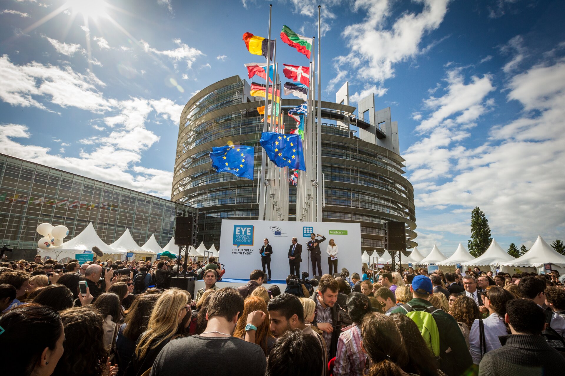 Na zdjęciu w tle widoczny gmach Parlamentu Europejskiego w Strasbourgu. Przed nim wysokie maszty i flagi państw Unii Europejskiej i samej Unii. Na pierwszym planie scena. Na scenie czworo ludzi. Wokół sceny tłumy ludzi.