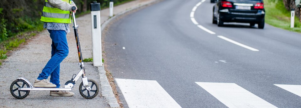 Kolorowe zdjęcie przedstawiające jezdnię z namalowanymi na niej pasami dla pieszych. Prawą stroną jezdni jedzie samochód, widoczny jest tylko jego odjeżdżający tył z czerwonymi światłami. Na chodniku, po lewej stronie jezdni, stoi dziecko z hulajnogą, widoczne od ramion w dół. Prawa noga oparta na hulajnodze. Dziecko ma założoną kamizelkę odblaskową.