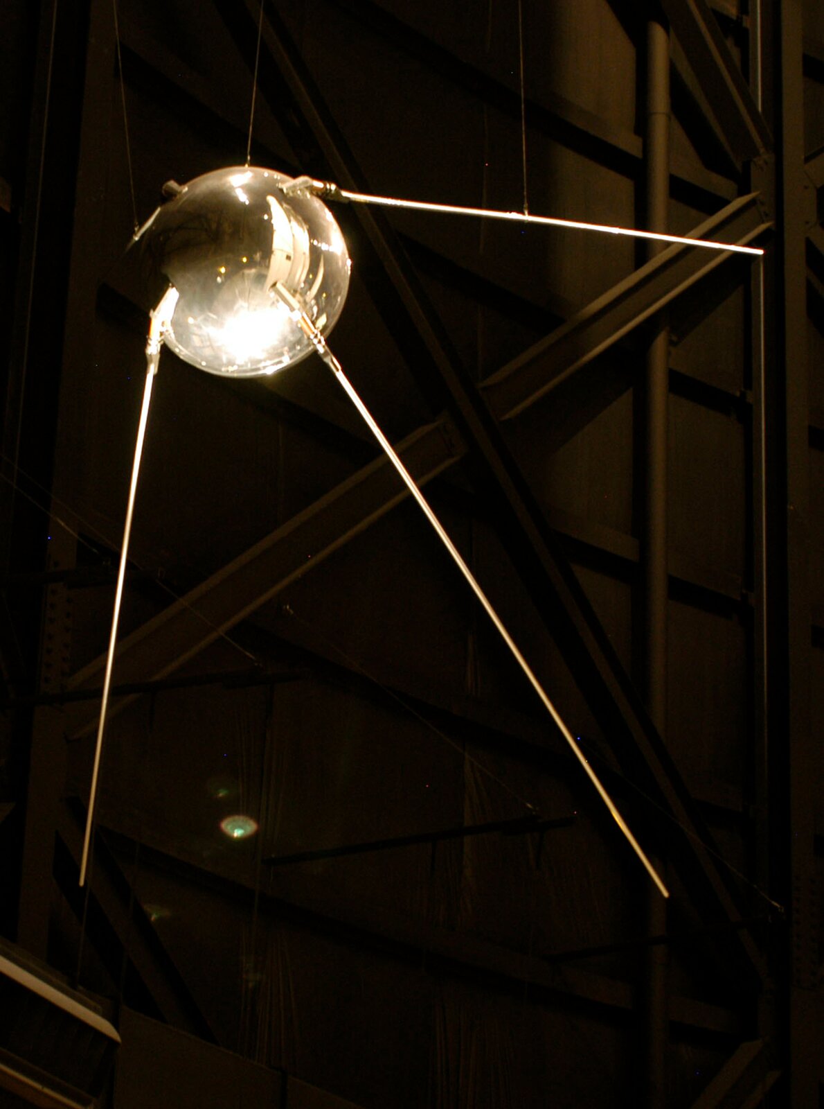 Zdjęcie przedstawia pierwszego sztucznego satelitę Ziemi – SPUTNIK 1. Tło ciemne, prawie czarne. Na zdjęciu widoczne urządzenie przypominające dużą, srebrną kulę z trzema cienkimi „nóżkami” odchodzącymi od niej. Powierzchnia błyszcząca.