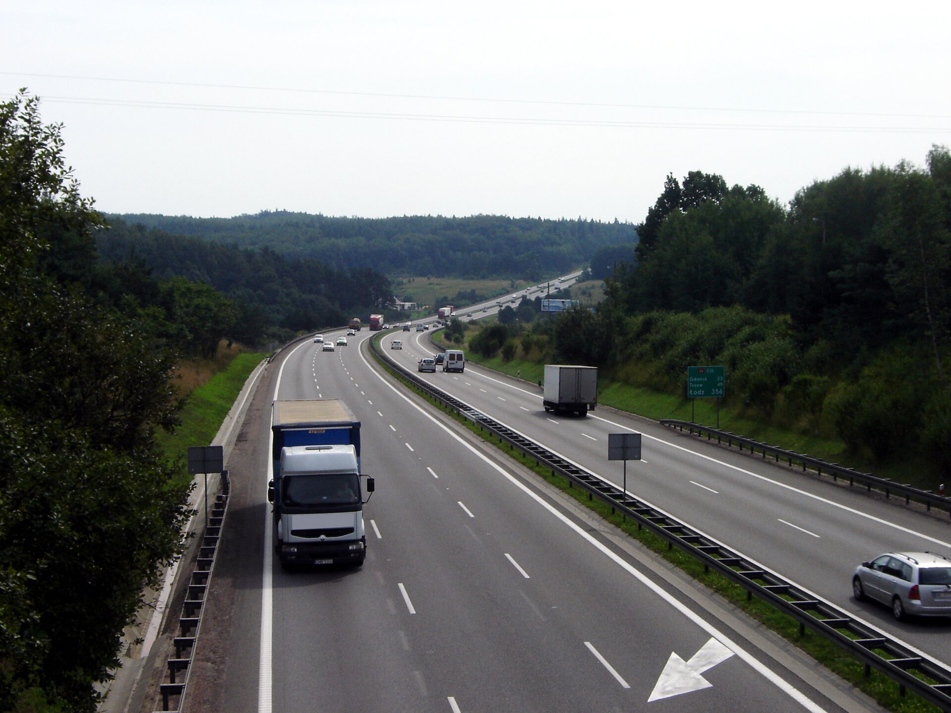 Fotografia prezentuje autostradę z trzema pasami ruchu w obu kierunkach. Autostradą jadą samochody ciężarowe i osobowe. Pobocza autostrady porośnięte drzewami, krzewami i trawą.