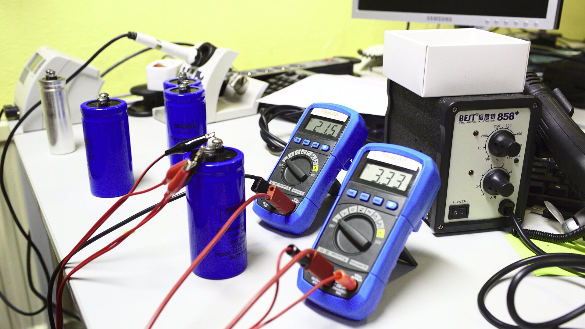 Zdjęcie przedstawia stół warsztatowy, którego użytkownik dokonuje operacji przywracania elementów elektronicznych zwanych kondensatorami, do pełnej sprawności. Kondensatory mają kształt dość dużych niebieskich cylindrów i stoją obok siebie w liczbie czterech sztuk. Do jednego z nich podłączone zostało źródło prądu oraz dwa multimetry mierzące napięcie przy obu zakończeniach, zwanych okładkami.