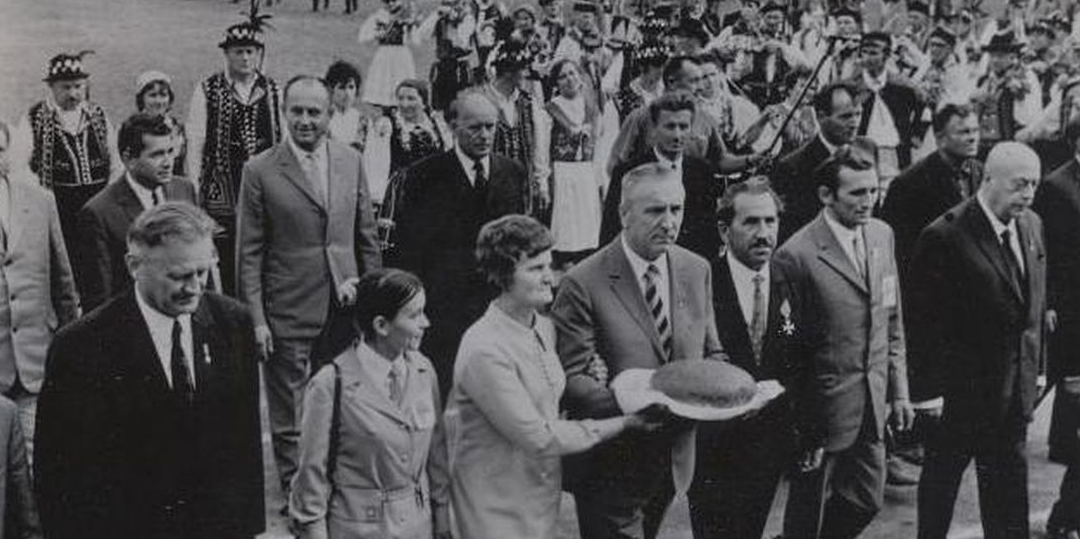 Fotografia czarno- biała przedstawiająca dożynki w czasach PRL. Edward Gierek, który przewodniczy pochodowi dożynkowemu otoczony tłumem współpracowników, działaczy niesie chleb.