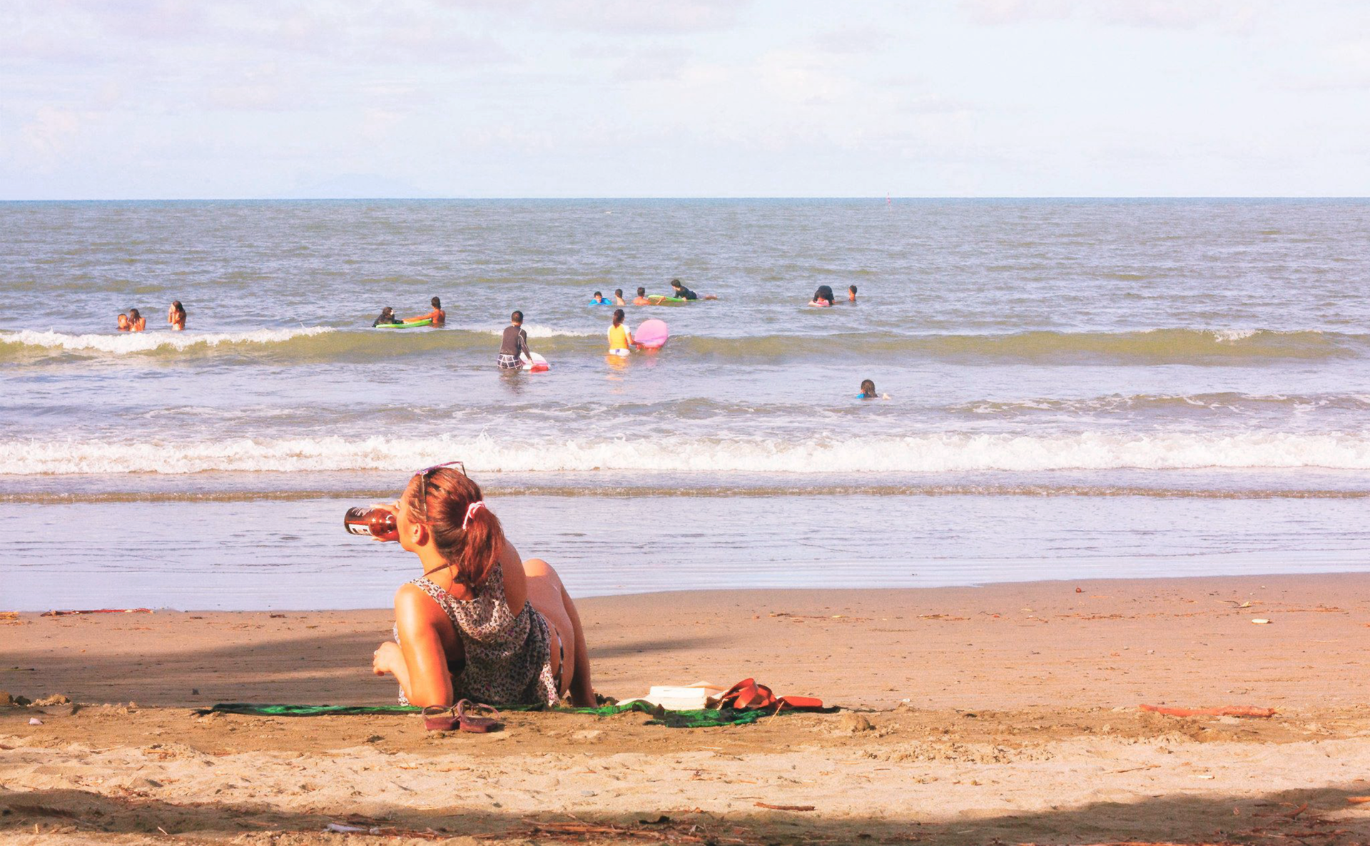 Kolorowe prostokątne zdjęcie przedstawia słoneczny letni dzień na plaży. Na pierwszym planie młoda kobieta leży na kocu, na piasku. Wsparta na prawym łokciu. W drugiej ręce trzyma butelkę z piwem. Pije piwo z butelki. W głębi morze. Małe fale. Kilkanaście osób pływa w morzu.