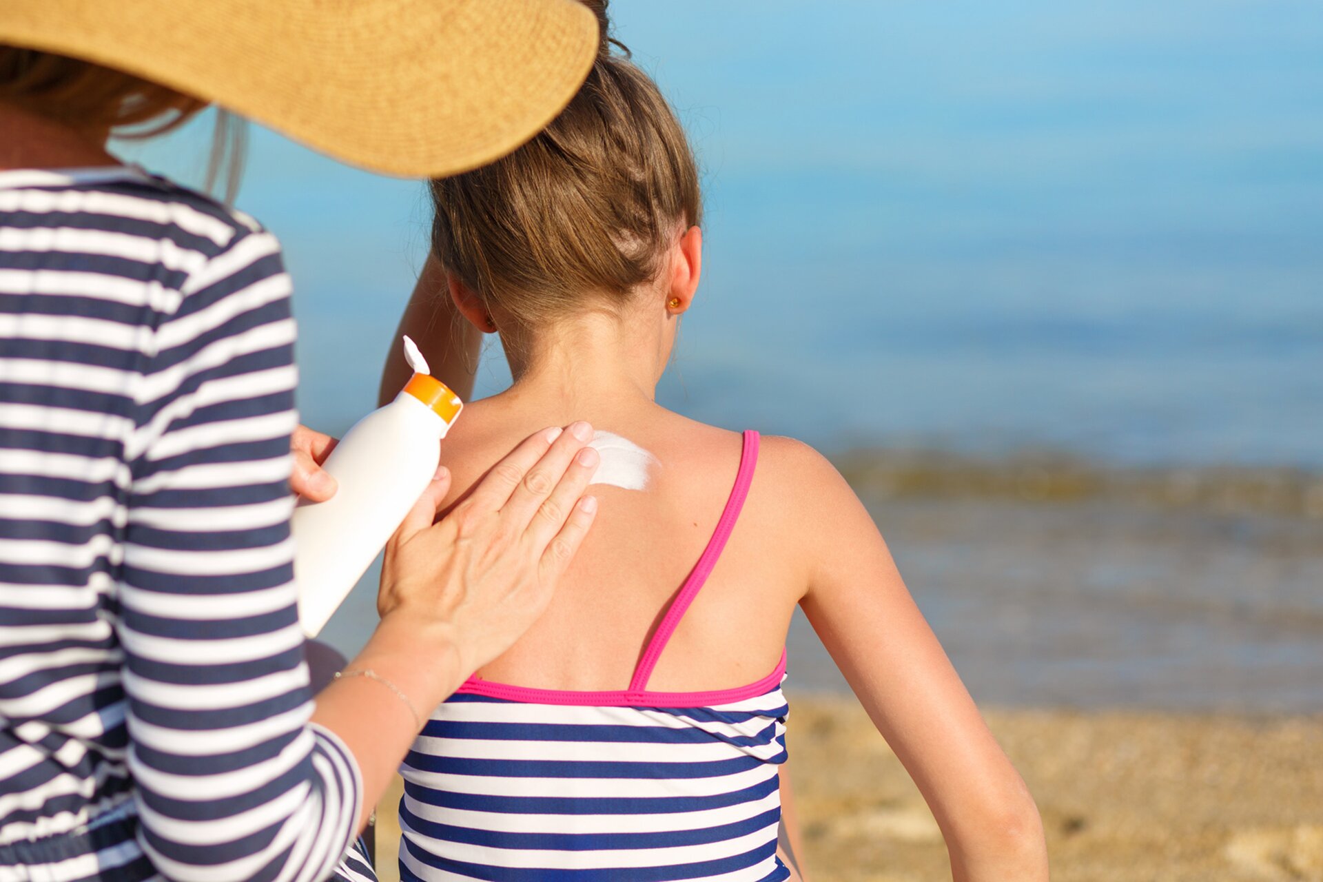 Fotografia prezentuje kobietę i dziewczynkę siedzące na plaży. Kobieta smaruje białym kremem plecy dziewczynki.