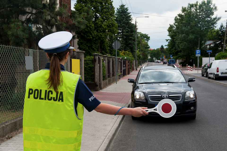 Zdjęcie przedstawia policjantkę zatrzymującą samochód do kontroli. Policjantka ustawiona jest tyłem do obserwatora zdjęcia, w wyciągniętej prawej ręce ma czerwony lizak. Zatrzymywany samochód to elegancka toyota w ciemnych barwach. Miejsce akcji to niezbyt ruchliwa droga w dzielnicy willowej.