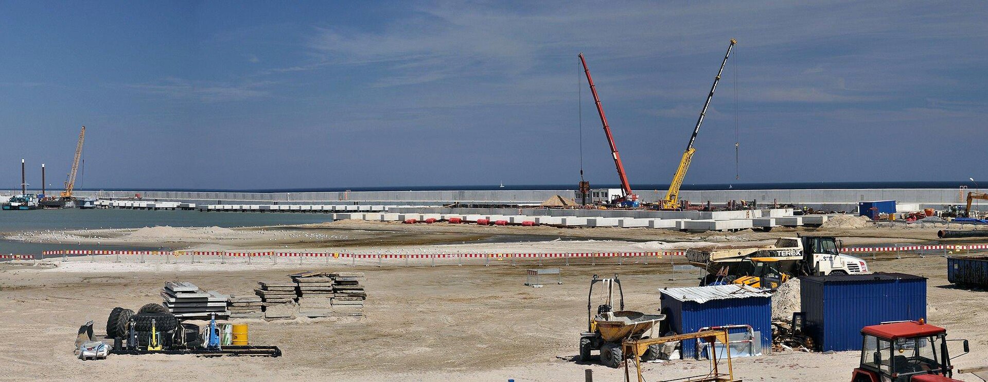 Kolorowe zdjęcie przedstawia gazoport w trakcie budowy. Słoneczny dzień. Plaża. Na plaży elementy budowy, urządzenia, samochody, koparki. Po prawej stronie, w głębi, dwa dźwigi. Na linii horyzontu betonowy element budowy w postaci długiego muru z płyt betonowych. W głębi zdjęcia ciemno niebieskie morze.