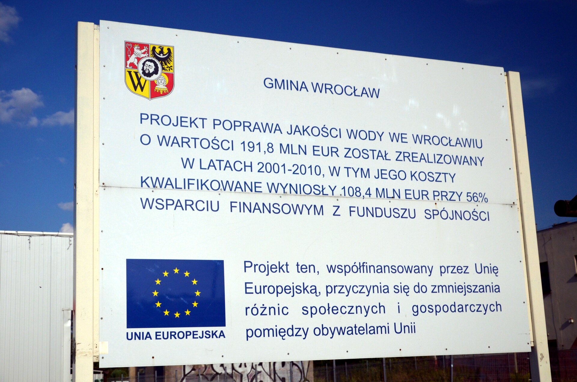 Na zdjęciu biała tablica z herbem Wrocławia i flagą Unii Europejskiej z napisem: Gmina Wrocław, projekt poprawa jakości wody we Wrocławiu o wartości sto dziewięćdziesiąt jeden i osiem dziesiątych miliona euro został zrealizowany w latach dwa tysiące jeden – dwa tysiące dziesięć, w tym jego koszty kwalifikowane wyniosły sto osiem i cztery dziesiąte miliona euro przy pięćdziesięcio sześcio procentowym wsparciu finansowym z funduszu spójności. Projekt ten, współfinansowany przez przez Unię Europejską, przyczynia się do zmniejszania różnic społecznych i gospodarczych pomiędzy obywatelami Unii.