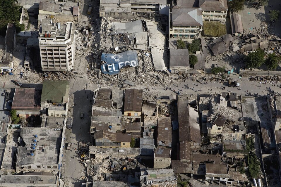 Zdjęcie przedstawia skutki trzęsienia ziemi oglądane z lotu ptaka. Słoneczny dzień, miasto Port-au-Prince, stolica Haiti w 2010 roku. Całe zdjęcie zajmują ruiny budynków mieszkalnych po trzęsieniu ziemi. W połowie zdjęcia, główna ulica sięga lewej i prawej krawędzi zdjęcia. Na ulicy mieszkańcy miasta. Wzdłuż ulicy, u dołu zdjęcia oraz z prawej strony również u góry, brązowe blaszane dachy ocalałych domów. Ulica częściowo zasypana gruzem zniszczonych budynków w centrum kadru. W dolnej połowie zdjęcia budynki są niskie. W górnej części zdjęcia ocalałe budynki to bloki mające po pięć lub sześć pięter. Ściany betonowych bloków białe bez widocznych pęknięć.