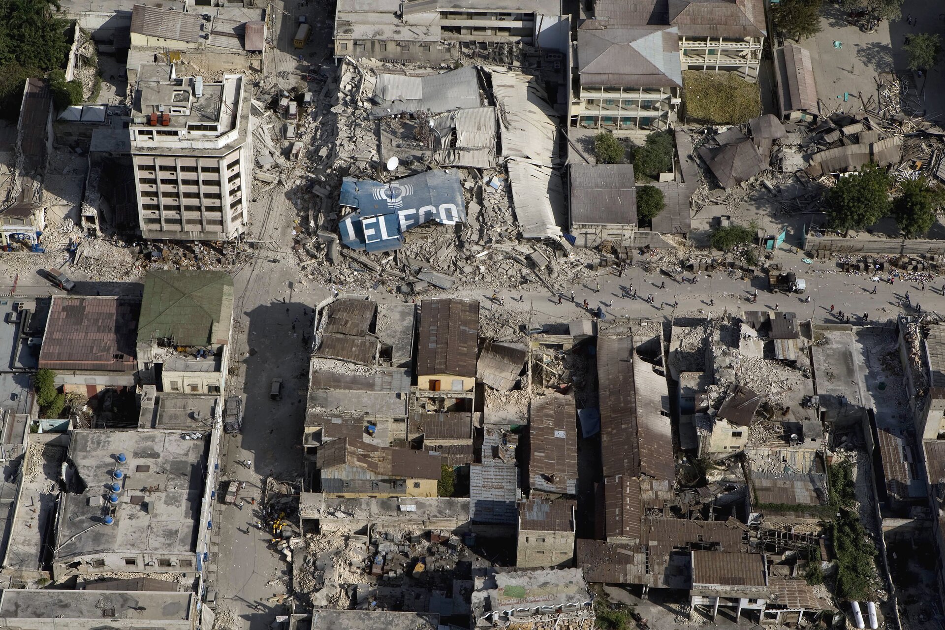 Zdjęcie przedstawia skutki trzęsienia ziemi oglądane z lotu ptaka. Słoneczny dzień, miasto Port-au-Prince, stolica Haiti w 2010 roku. Całe zdjęcie zajmują ruiny budynków mieszkalnych po trzęsieniu ziemi. W połowie zdjęcia, główna ulica sięga lewej i prawej krawędzi zdjęcia. Na ulicy mieszkańcy miasta. Wzdłuż ulicy, u dołu zdjęcia oraz z prawej strony również u góry, brązowe blaszane dachy ocalałych domów. Ulica częściowo zasypana gruzem zniszczonych budynków w centrum kadru. W dolnej połowie zdjęcia budynki są niskie. W górnej części zdjęcia ocalałe budynki to bloki mające po pięć lub sześć pięter. Ściany betonowych bloków białe bez widocznych pęknięć.