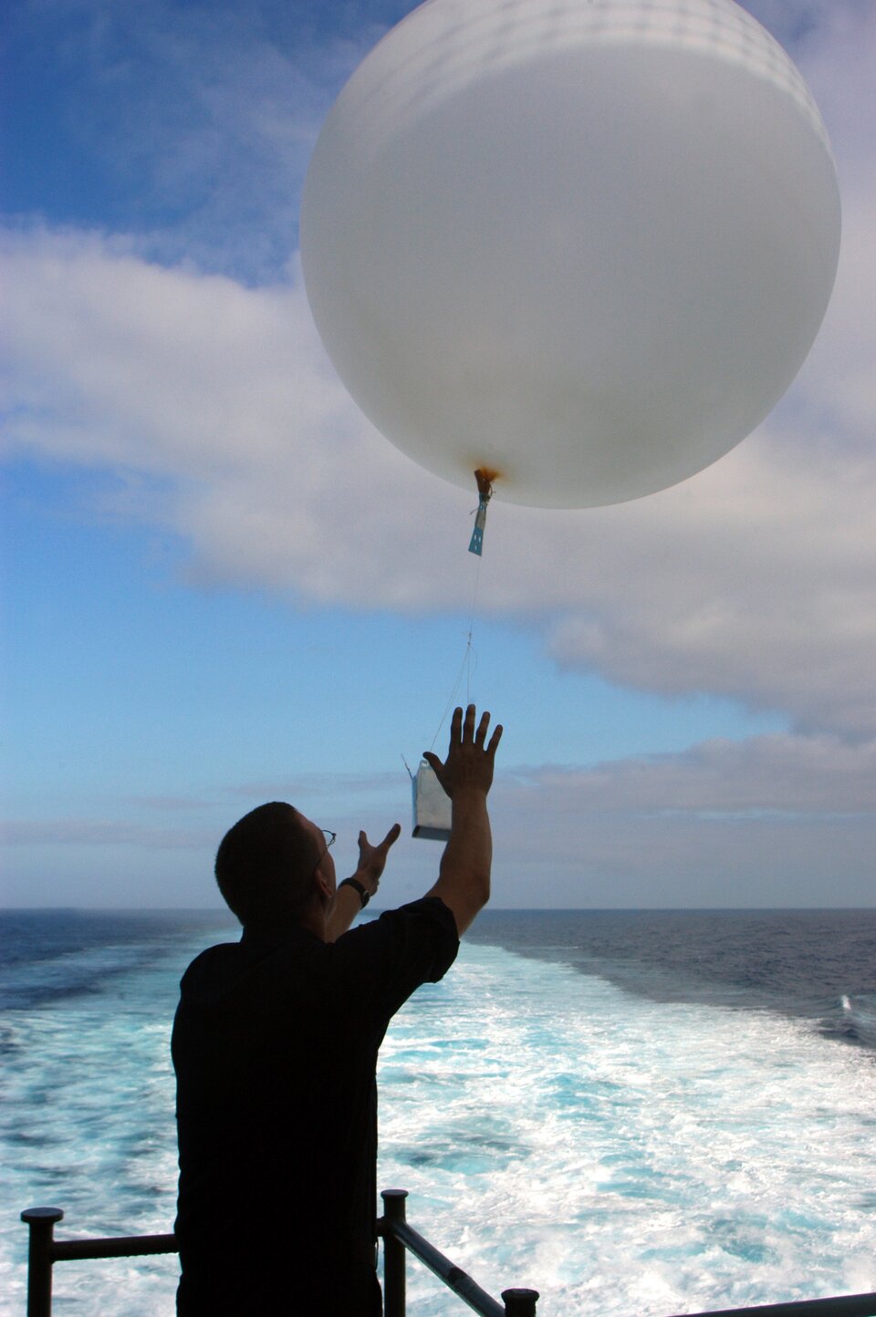 Zdjęcie przedstawia balon meteorologiczny. Niewielki balon ma kształt kuli o średnicy około dwóch metrów. Pod balonem podwieszona jest skrzynka z przyrządami służącymi do pomiaru składników pogody.