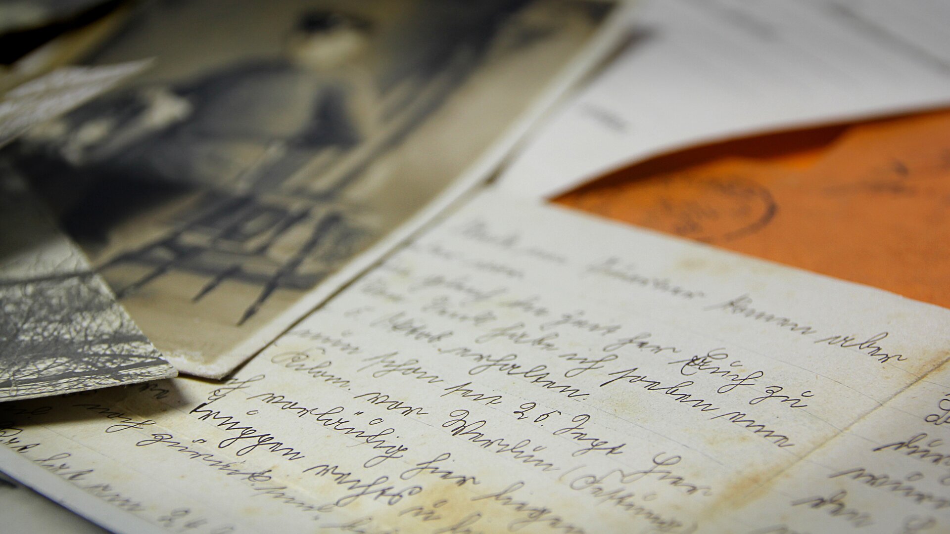 Zdjęcie przedstawia list napisany ręcznie. Papier jest nieco pożółkły, co może świadczyć o tym, że jest to list sprzed wielu lat. Obok niego leży czarno-biała fotografia. Widać na niej zarys człowieka. Stoi wyprostowany. Prawdopodobnie ma na sobie mundur.