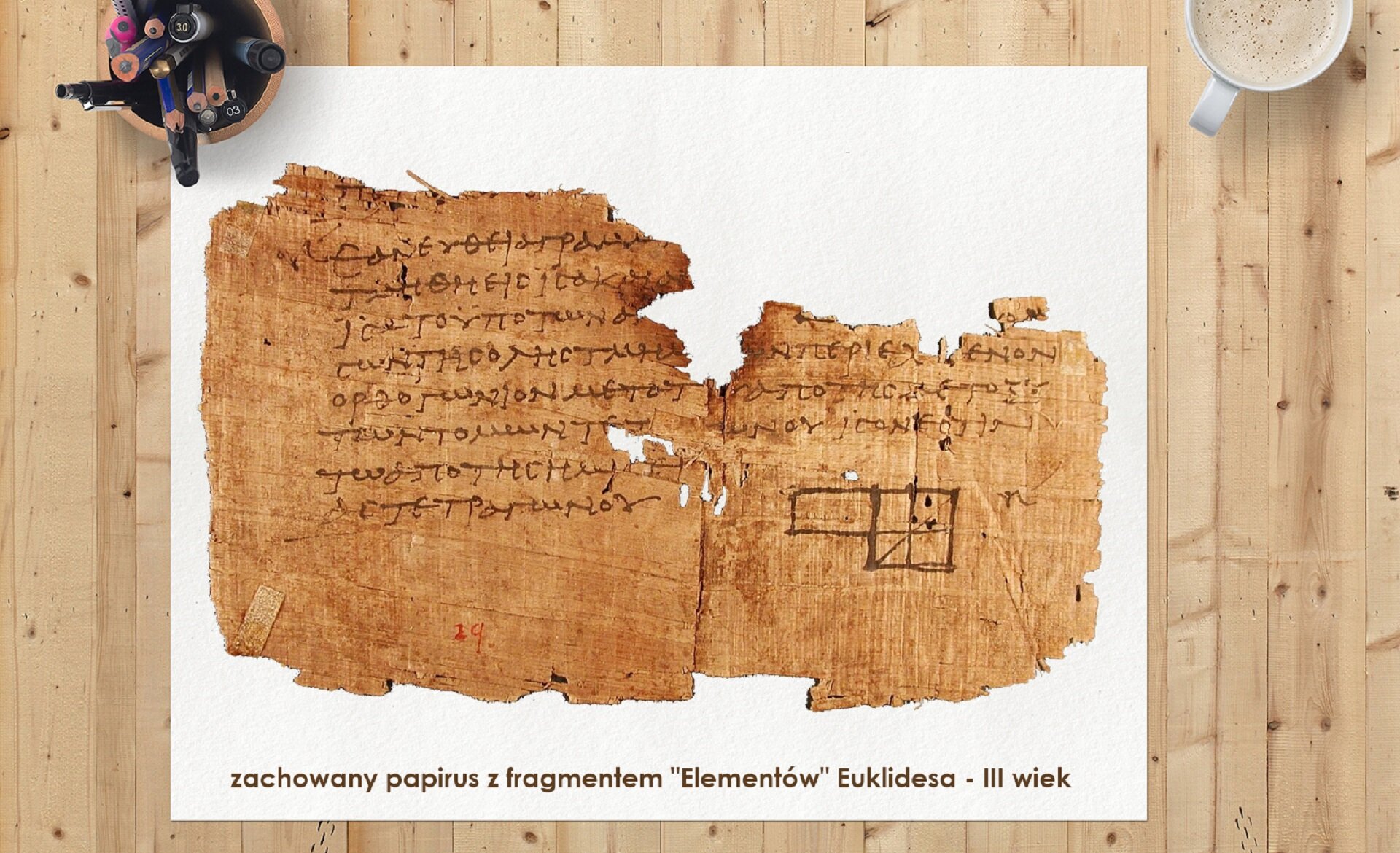 Ilustracja przedstawia sfotografowaną z góry drewnianą płaszczyznę wykonaną z jasnych desek. Na deskach leży biały arkusz. Na nim leży fragment wystrzępionego papirusu. Papirus pokryty jest pismem greckim. U dołu tekstu widać rysunki czworoboków. Na dole białego arkusza widnieje napis informujący czym jest pokazany papirus : „zachowany papirus z fragmentem „Elementów” Euklidesa – III wiek” Lewy górny róg białego arkusza dociśnięty jest do blatu kubkiem, w którym znajdują się kolorowe kredki, długopisy i pisaki. W prawym górnym rogu widać fragment filiżanki z kawą.