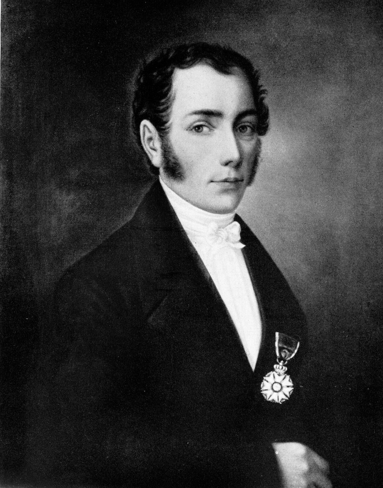 Zdjęcie przedstawia Josepha von Fraunhofera. Zdjęcie czarno białe. Mężczyzna w wieku około trzydziestu lat. Tors zwrócony w prawo. Twarz zwrócona w stronę obserwatora. Twarz pociągła. Skóra jasna gładko ogolona. Czoło wysokie. Czarne proste włosy zaczesane do tyłu. Uszy duże. Bokobrody długie i czarne. Brwi czarne wąskie. Nos długi wąski. Ostro zakończony podbródek. Wysoki kołnierz białej koszuli zakrywa szyję. Mężczyzna ubrany w czarną marynarkę. Na lewej klapie marynarki duży order. Okrągły order zawieszony na wstążce.