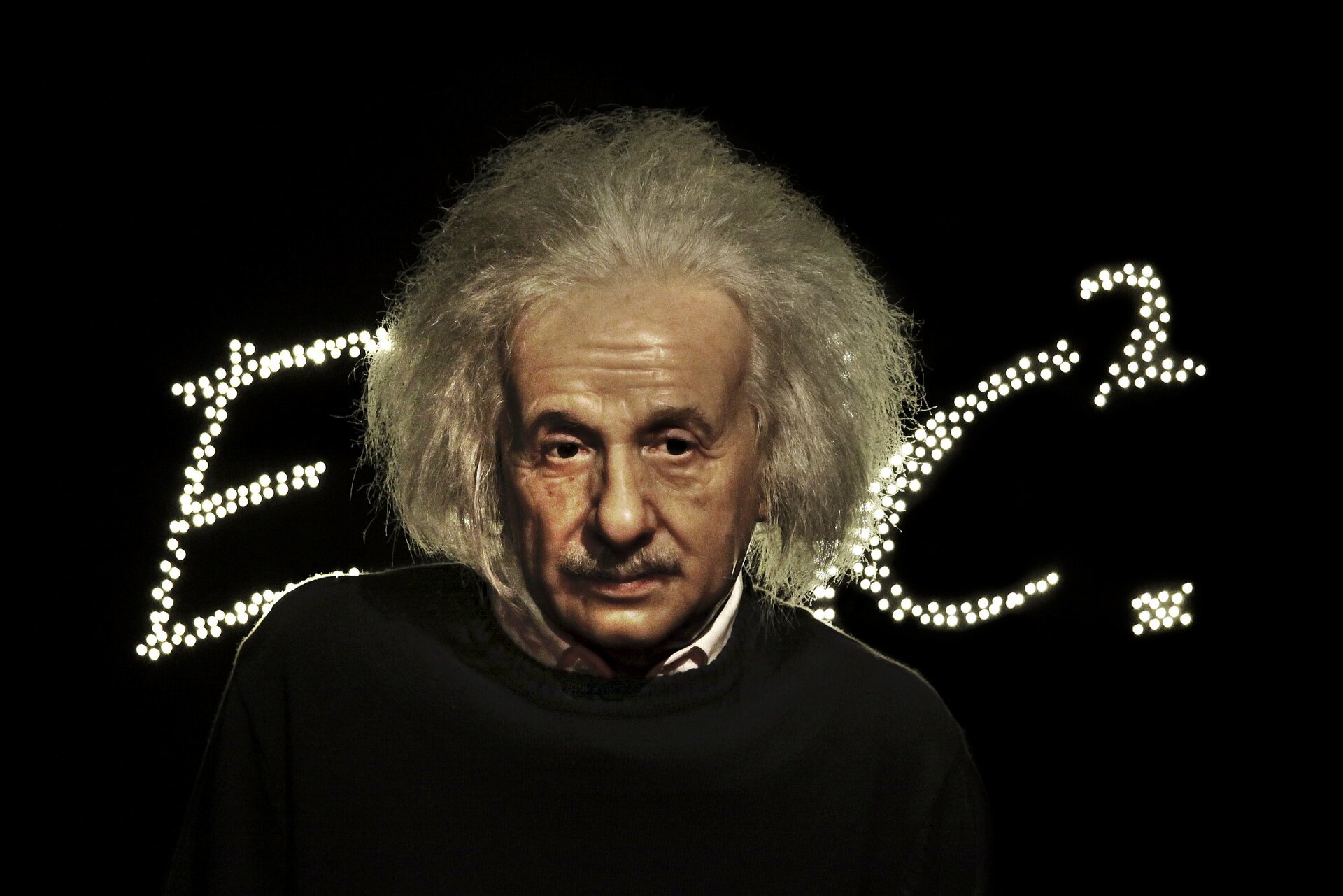Zdjęcie przedstawia podobiznę Alberta Einsteina, a dokładniej mówiąc przedstawiającą go figurę w gabinecie figur naturalnych Madame Tussaud na tle ułożonego z licznych punktowych świateł wzoru E równa się m c kwadrat. Uczony przedstawiony w wieku średnim ukazany jest od ramion w górę, spogląda nieco w bok z zamyślonym wyrazem twarzy.