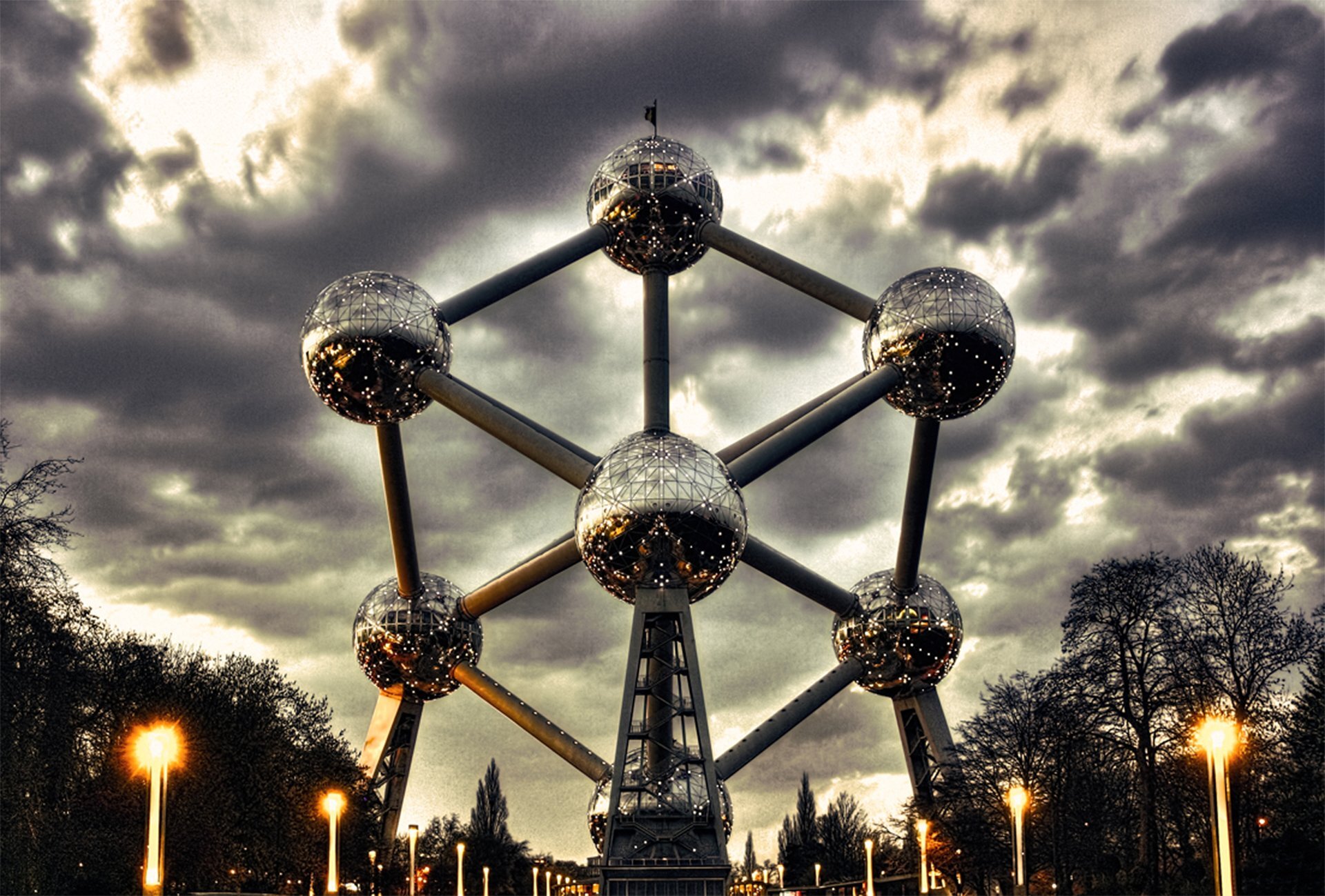 Zdjęcie przedstawia budowlę Atomium składającą się z dziewięciu metalowych kul połączonych ze sobą, z czego na zdjęciu widocznych jest siedem. Zdjęcie wykonano wieczorową porą, przez co od kul odbijają się liczne światła miasta. Na tle Atomium rozpościera się niebo pokryte burzowymi chmurami, poniżej znajdują się drzewa i latarnie uliczne.
