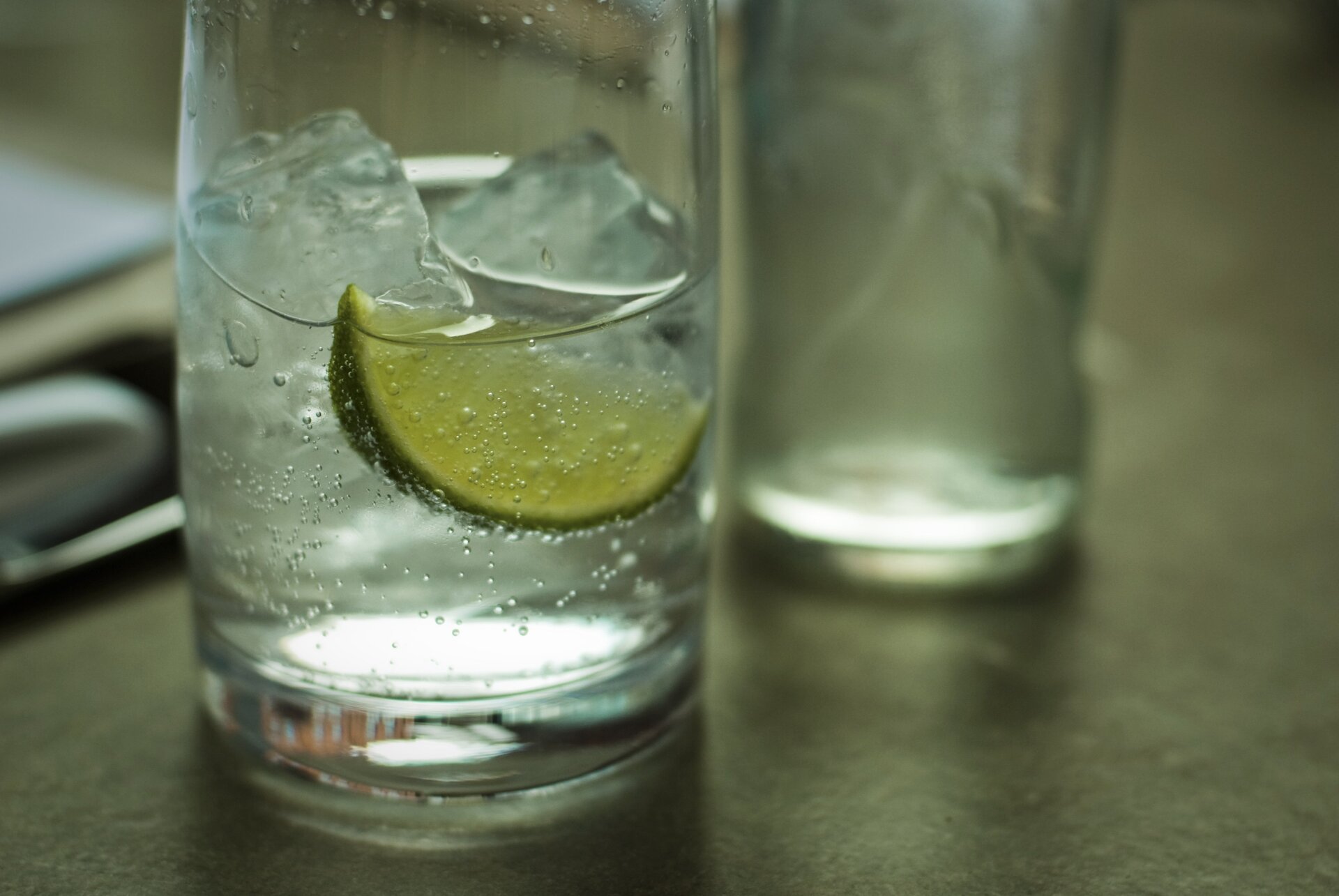 Zdjęcie przedstawia stojącą na stole szklankę do połowy napełnioną wodą mineralną i pływającymi w niej kostkami lodu i plastrem limonki. W tle zdjęcia w obszarze nieostrości znajdują się pusta szklanka i inne nierozpoznawalne przedmioty.
