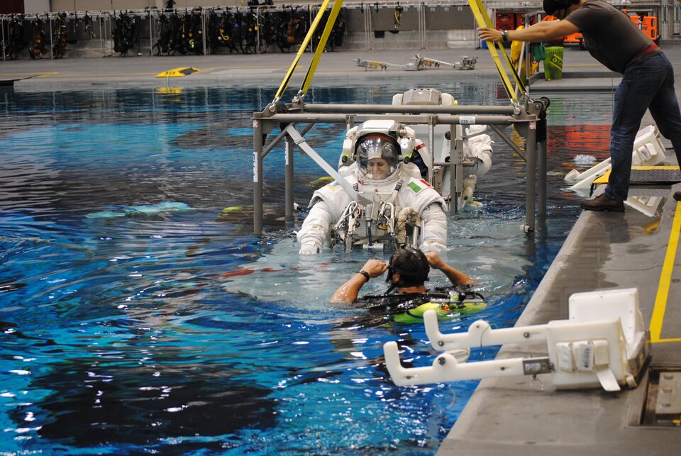 Zdjęcie przedstawia astronautkę podczas szkolenia. Astronautka ubrana jest w skafander. Ćwiczenia odbywają się w basenie. Astronautka zanurzona jest do połowy. W basenie znajduje się również mężczyzna. Mężczyzna wyposażony jest w sprzęto do nurkowania.