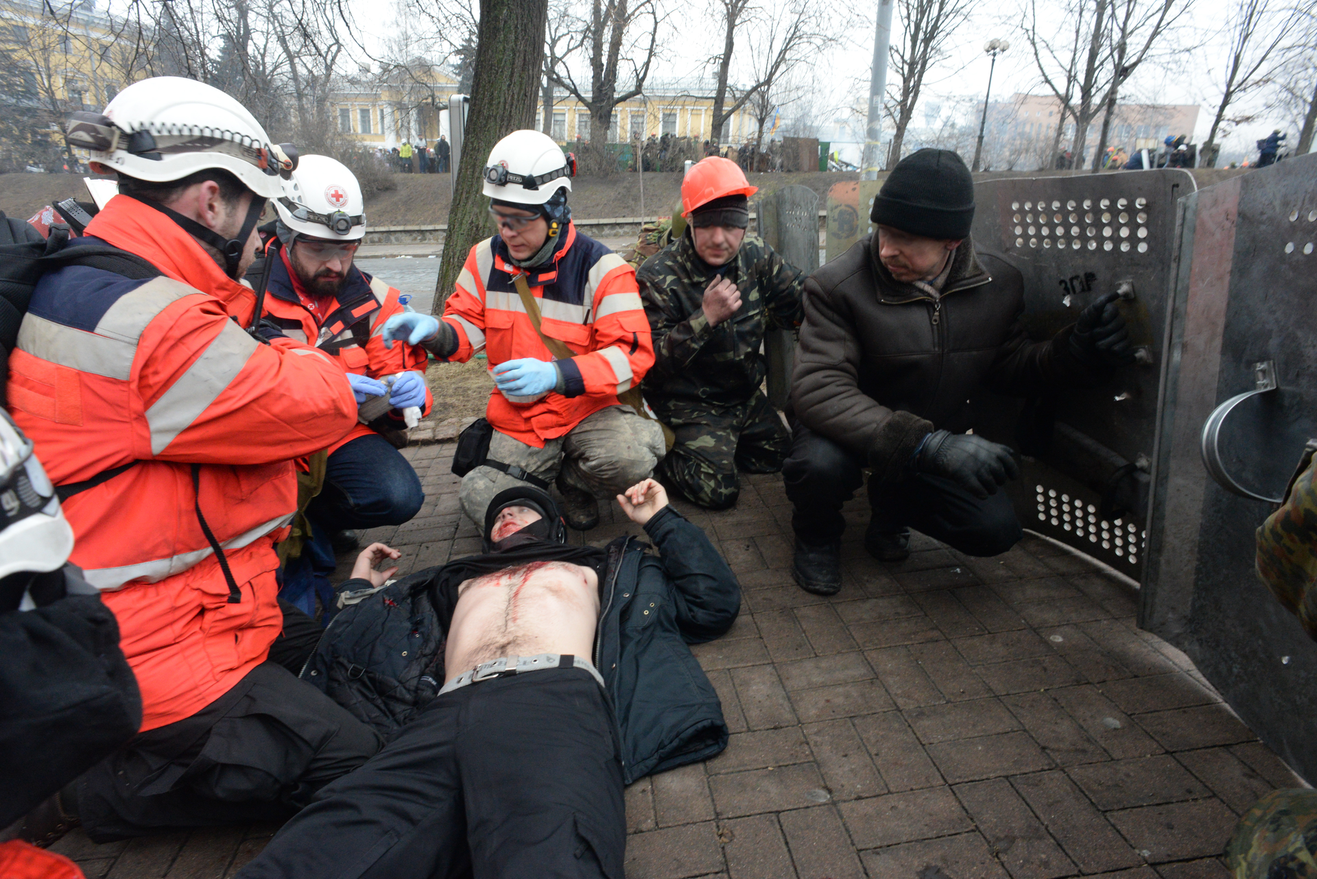 Kolorowe zdjęcie przedstawia trzech ratowników udzielających pomocy rannej osobie. Ratowicy znajdują się na chodniku. Ratownicy w lekkim przysiadzie po lewej stronie zdjęcia. Ubrani w czerwone kurtki z odblaskowymi pasami wokół ramion. Poziome pasy powyżej klatki piersiowej oraz na plecach. Na głowach białe kaski. Na kaskach umocowane latarki. Na dłoniach gumowe rękawice ochronne. Na prawo od ratowników leży osoba poszkodowana z odkrytym brzuchem i klatką piersiową. W tle dwie osoby w przysiadzie. Po prawej stronie zdjęcia płotek.