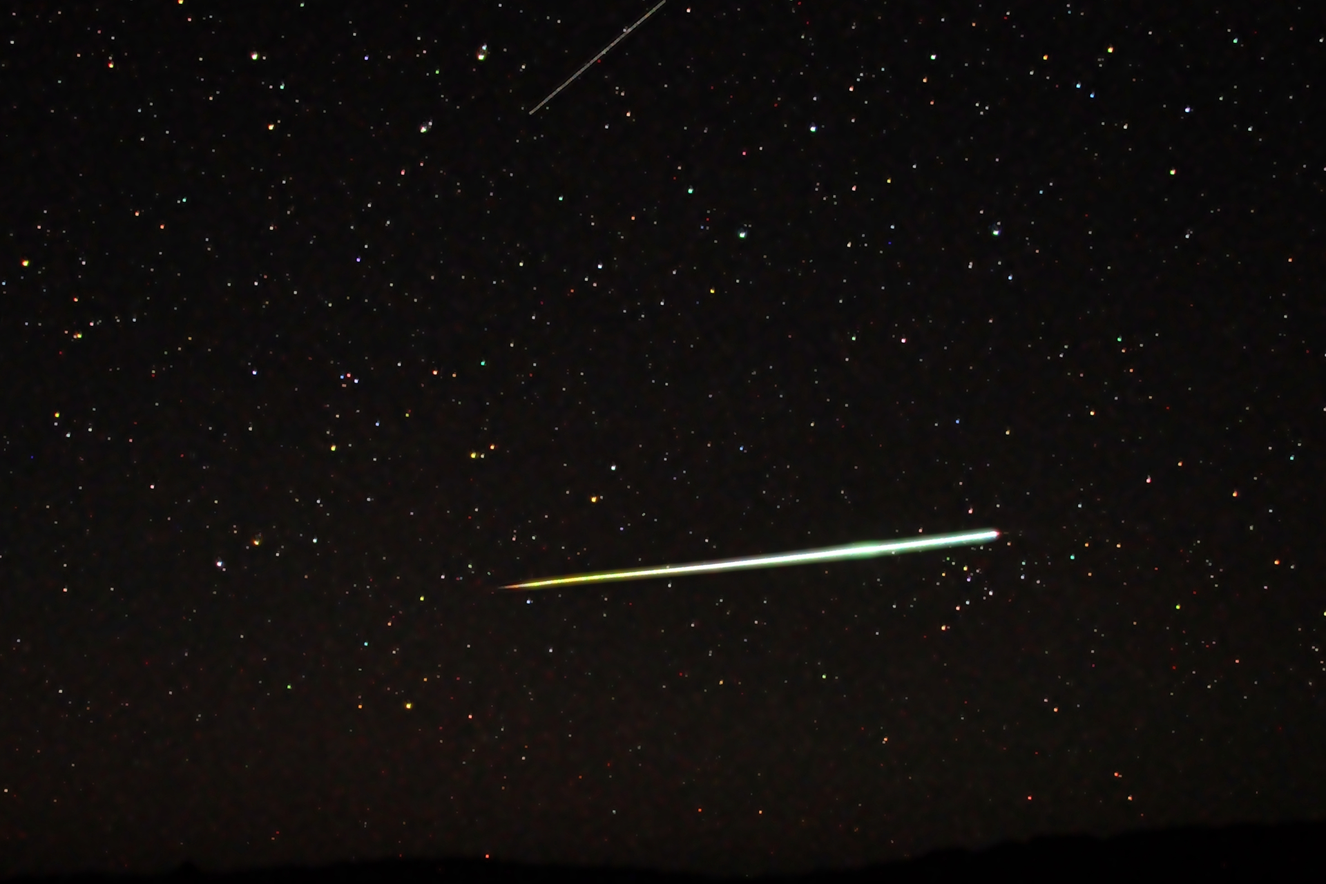 Fotografia prezentuje meteor. Na czarnym niebie widoczne gwiazdy w postaci białych licznych punkty różnej wielkości. Na środku fotografii widoczna biała wąska smuga.