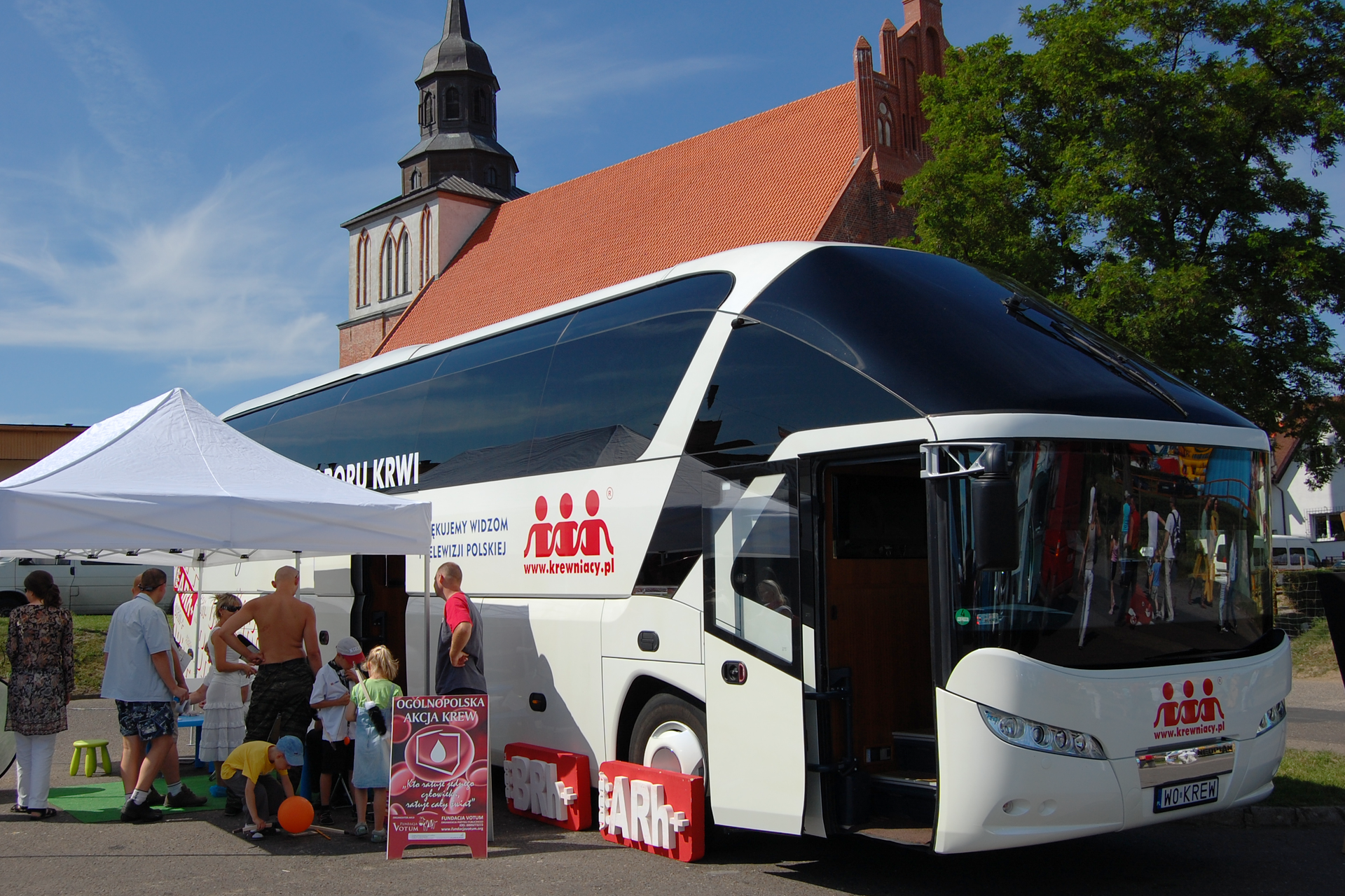 Akcja zbierania krwi. Na zdjęciu widać specjalnie wyposażony do pobierania krwi autobus-ambulans z namiotem do rejestracji. Pod namiotem stoją dorośli zapisując się do oddania krwi. Obok dzieci bawią się balonem. Autobus stoi pod kościołem na ulicy.