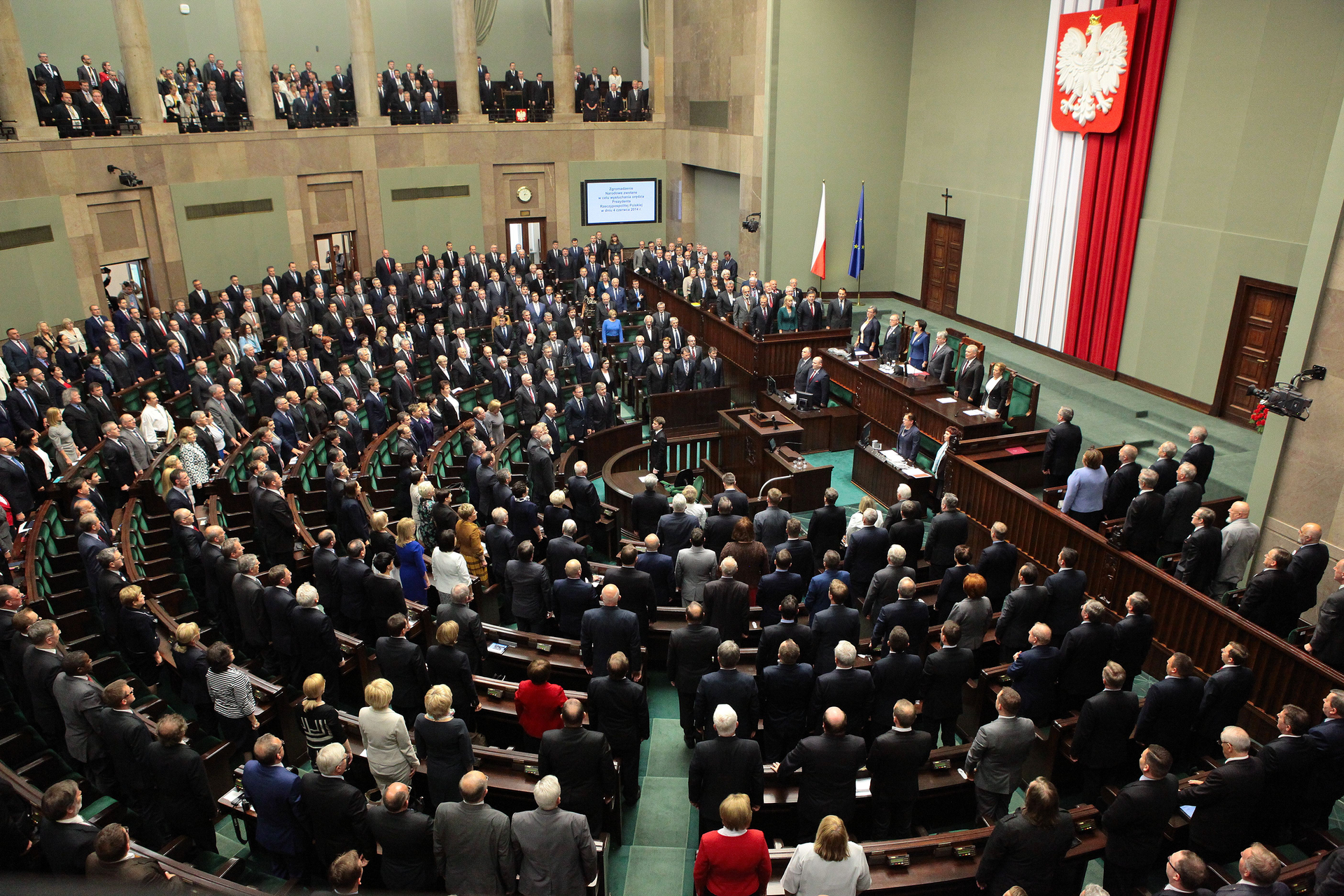Kolorowe zdjęcie przedstawia salę Sejmu RP. Zdjęcie z góry. Sala w kształcie półokręgu. Po prawej stronie wysoka ściana. Na środku, w pionie, zawieszony materiały z zakładkami. Po lewej stronie kolor biały, po prawej kolor czerwony. Na materiale zawieszone duże godło Polski. Biały orzeł w koronie na czerwonym tle. Przed ścianą z godłem długi rząd ławek równolegle do ściany. Pozostała część sali w kształcie półokręgu. Siedzenia ułożone w formie ławek, w kształcie półokręgów. Osoby biorące udział w posiedzeniu stoją przed swoimi krzesłami. W centralnej części sali, w środku półokręgu, mównica. Wokół sali balkon wsparty na kolumnach. Na balkonie obserwatorzy posiedzenia Sejmu.