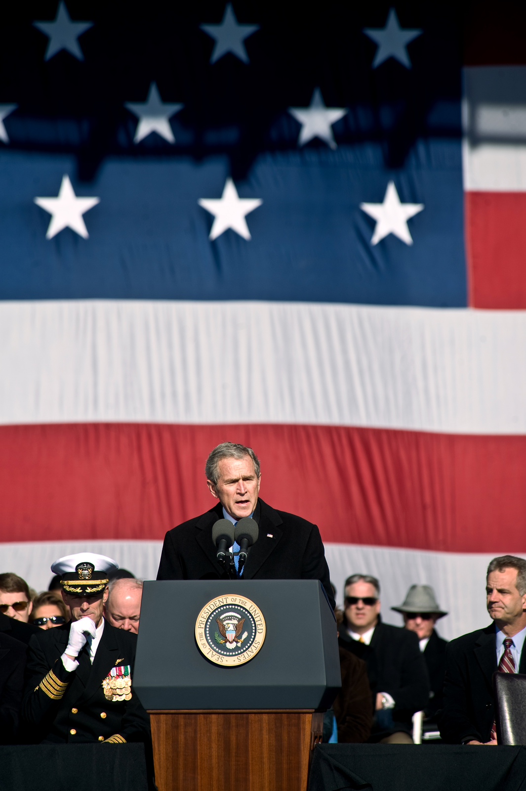 Zdjęcie przedstawia George`a Busha w trakcie przemówienia. Prezydent Bush stoi za mównicą. Ubrany w czarny płaszcz wygłasza przemówienie. W tle siedzą politycy, wojskowi. Wszyscy ubrani na czarno. Niektóre osoby mają czarne okulary. Na ścianie, w głębi zdjęcia, rozwieszona duża flaga amerykańska. Biało czerwone poziome pasy. Powyżej niebieski prostokąt z białymi pięcioramiennymi gwiazdami.