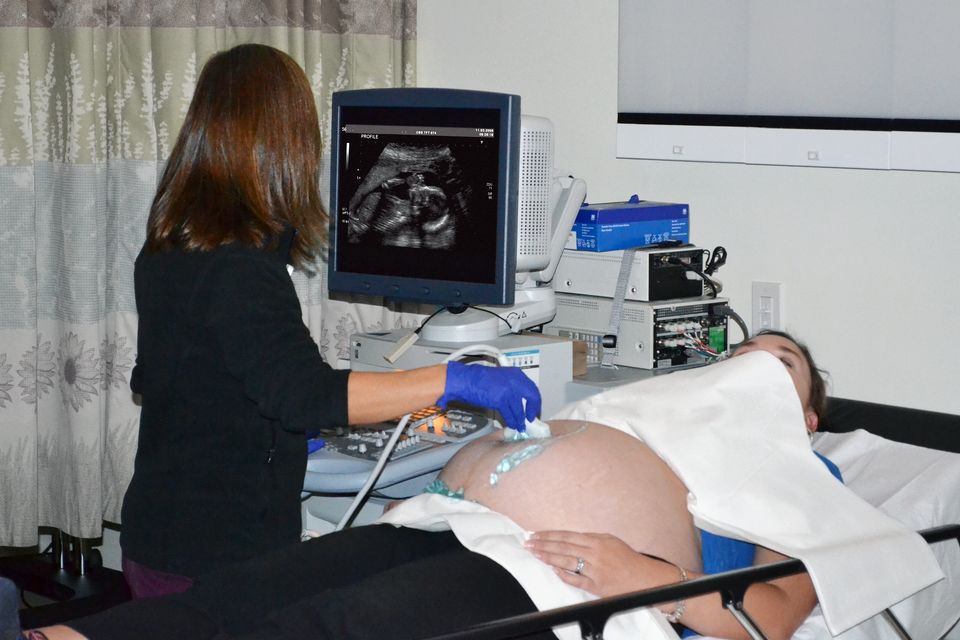Fotografia przedstawia lekarza w trakcie badania ultrasonograficznego. Po odsłoniętym brzuchu kobiety w zaawansowanej ciąży lekarka przesuwa głowicę aparatu, obserwując obraz dziecka na monitorze.