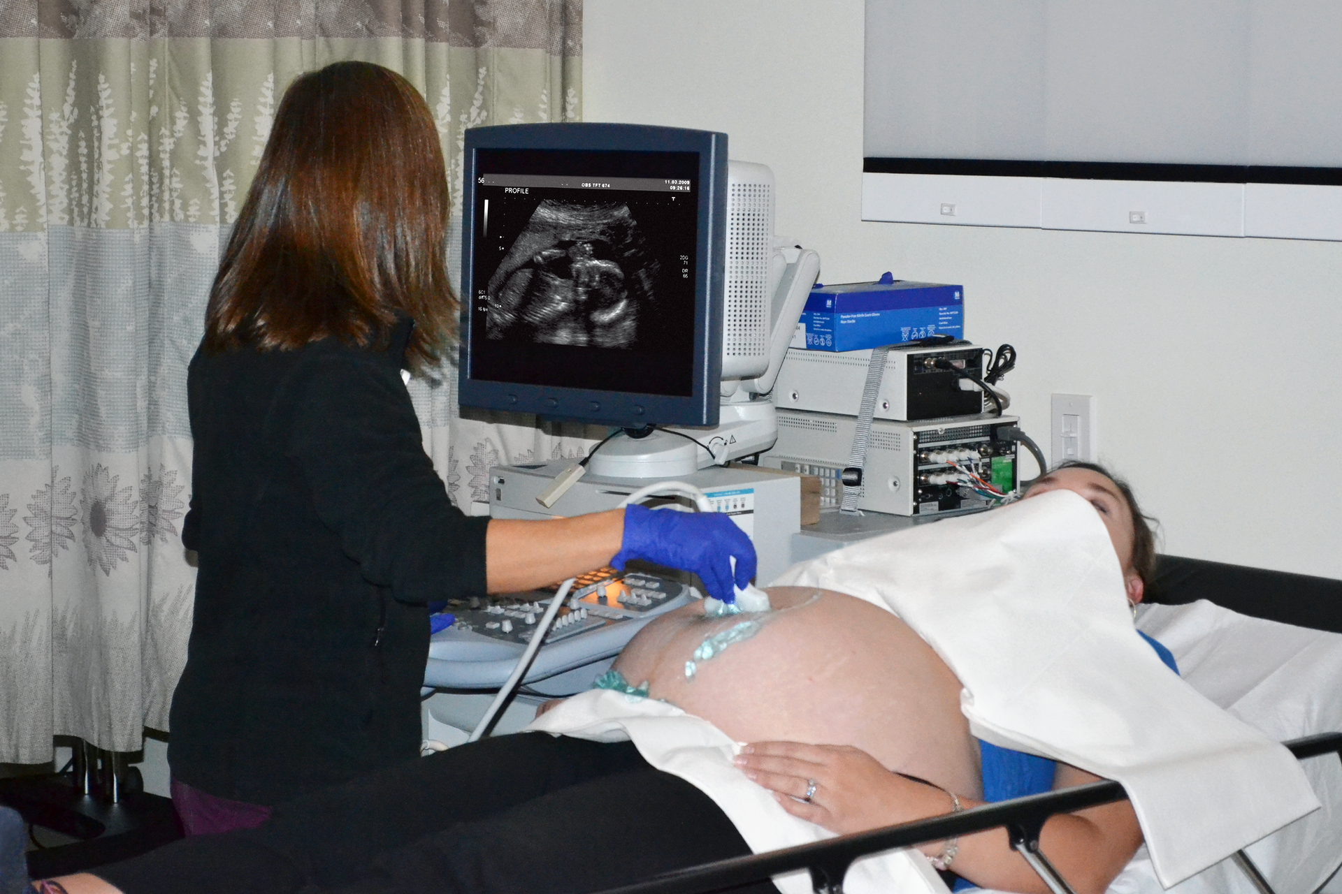 Fotografia przedstawia lekarza w trakcie badania ultrasonograficznego. Po odsłoniętym brzuchu kobiety w zaawansowanej ciąży lekarka przesuwa głowicę aparatu, obserwując obraz dziecka na monitorze.