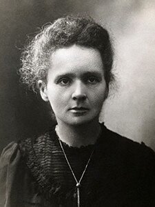 Czarno-białe zdjęcie portretowe Marii Skłodowskiej-Curie. Kobieta zwrócona twarzą w kierunku obserwatora. Ma długie włosy w kolorze ciemny blond. Fryzura lekko pofalowana, zaczesana do tyłu. Wysokie czoło. Brwi ciemne, gęste. Oczy duże. Nos mały. Usta wąskie. Twarz pociągła, bez zmarszczek. Uszy małe. Kobieta ubrana w czarną sukienkę. Wokół dekoltu ozdobne drobne marszczenia materiału.